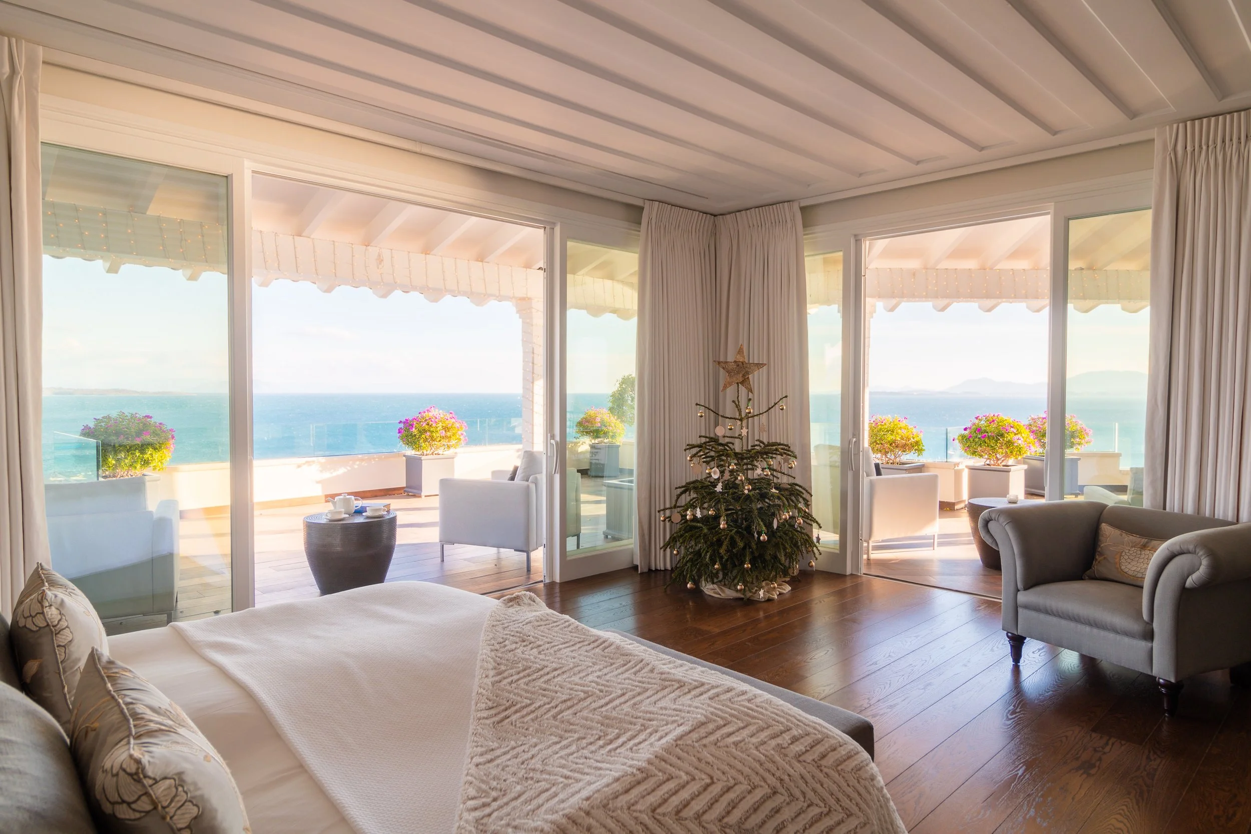 A living room with a view of the ocean. There's a Christmas tree in the center, and large sliding glass doors lead to a balcony with outdoor seating and potted flowers.