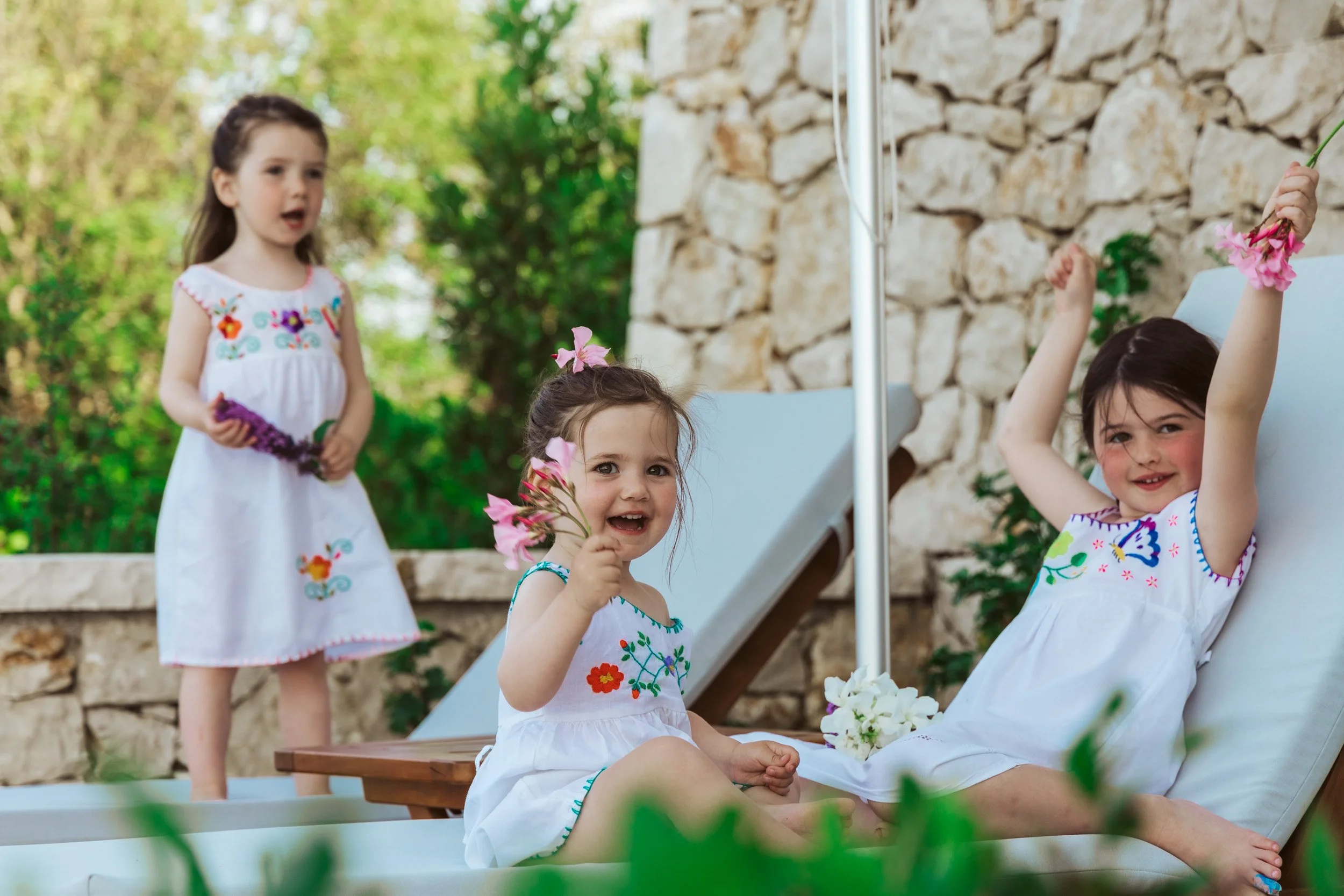 Three young girls in white dresses with colorful embroidery, two sitting on a lounge chair and one standing behind them outdoors, holding flowers and smiling, with a stone wall and green foliage in the background.