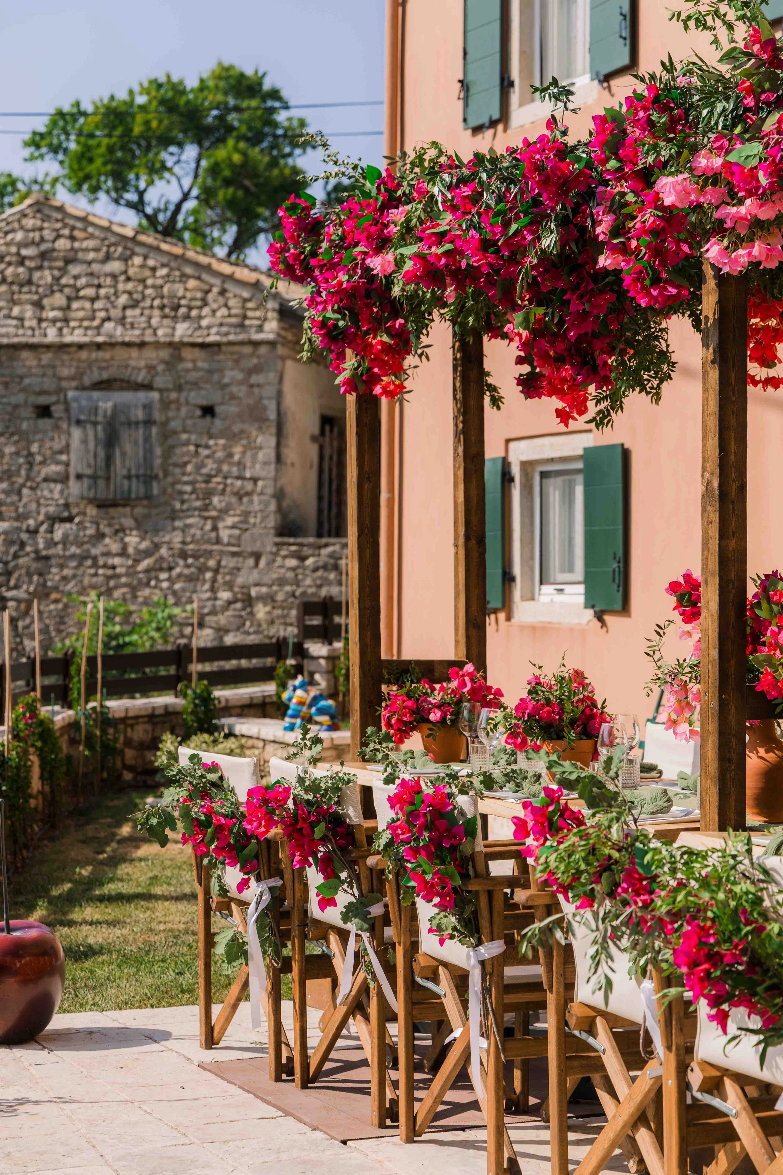 Outdoor dining setup with a long table and wooden chairs decorated with pink and red flowers, under a wooden frame with hanging flower baskets, next to a peach-colored building with window shutters, in a garden with an old stone building and trees in
