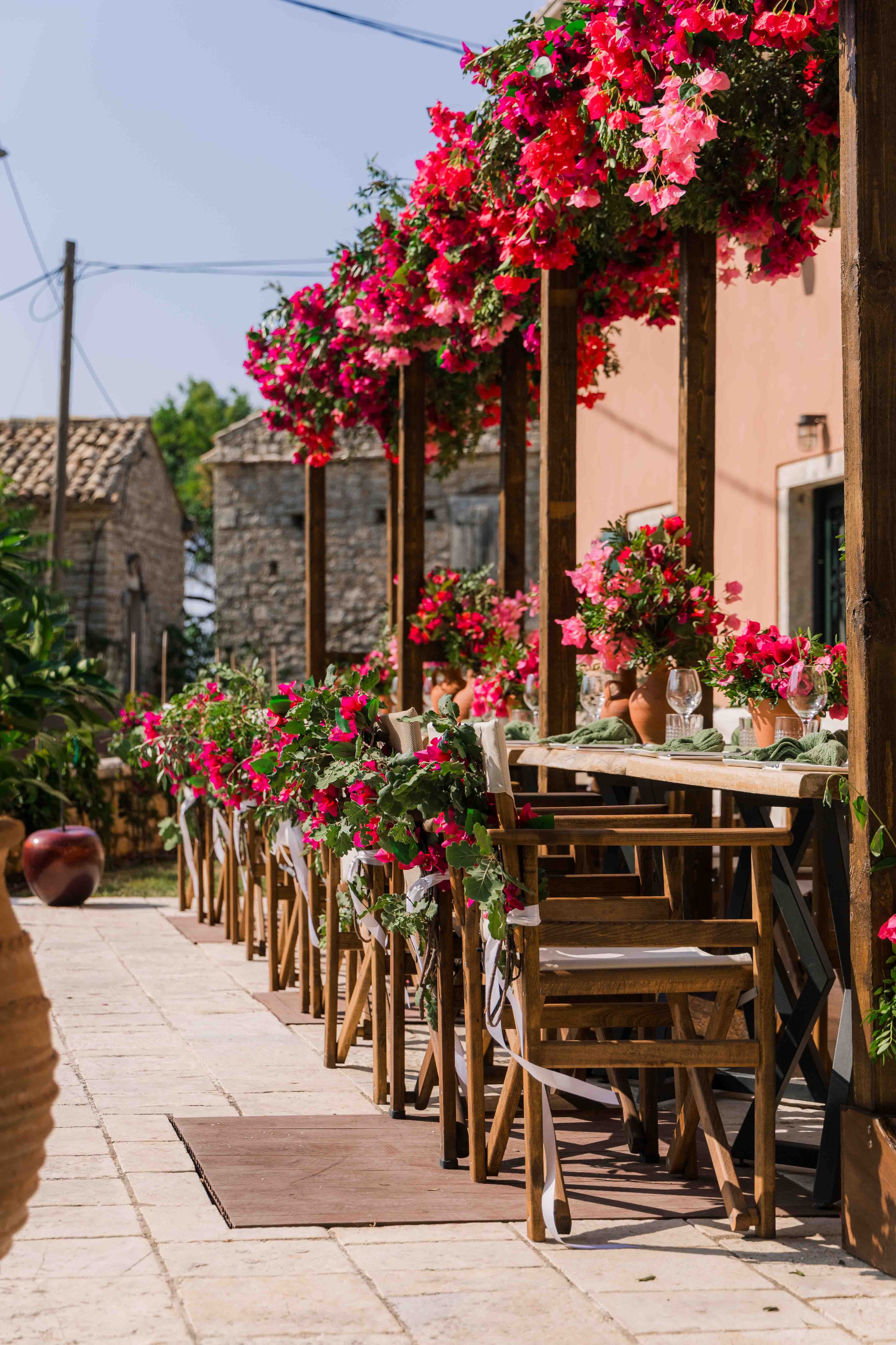 An outdoor patio decorated with pink and red flowers, including bougainvillea, along a wooden pergola with tables set for dining and surrounded by stone buildings.