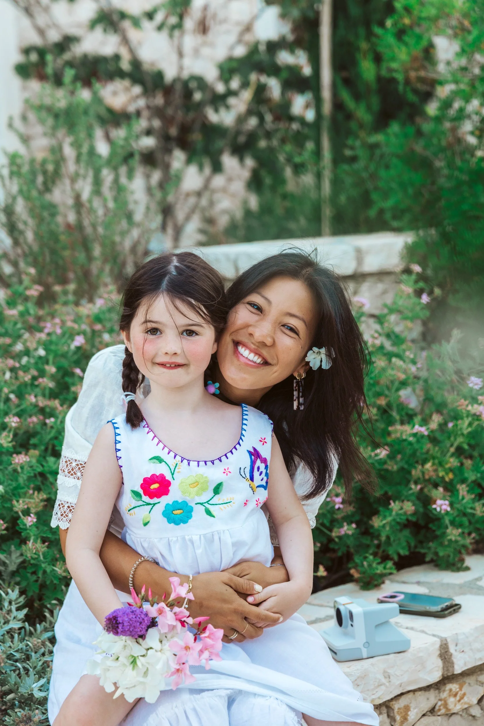 A woman with dark hair and a flower in her hair sitting outdoors on a stone bench, hugging a young girl with braided hair, both smiling; the girl is holding a bouquet of pink and purple flowers, and they are surrounded by greenery and pink flowers.