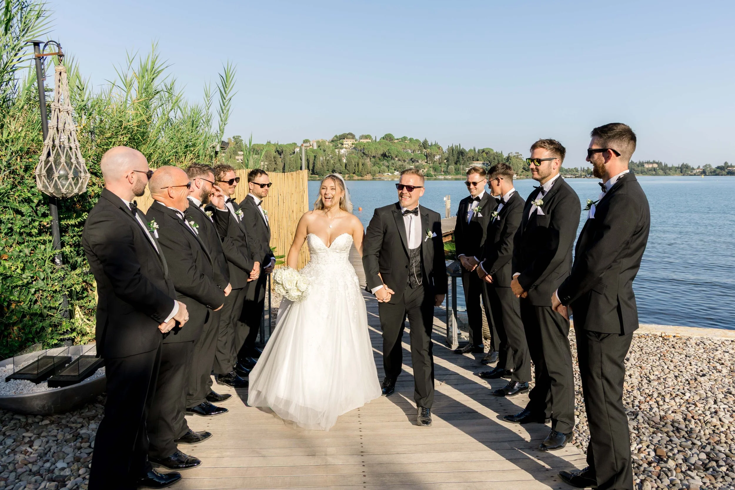 Bride and groom walking down the aisle by a lake with groomsmen and bridesmaids on either side during a wedding ceremony.