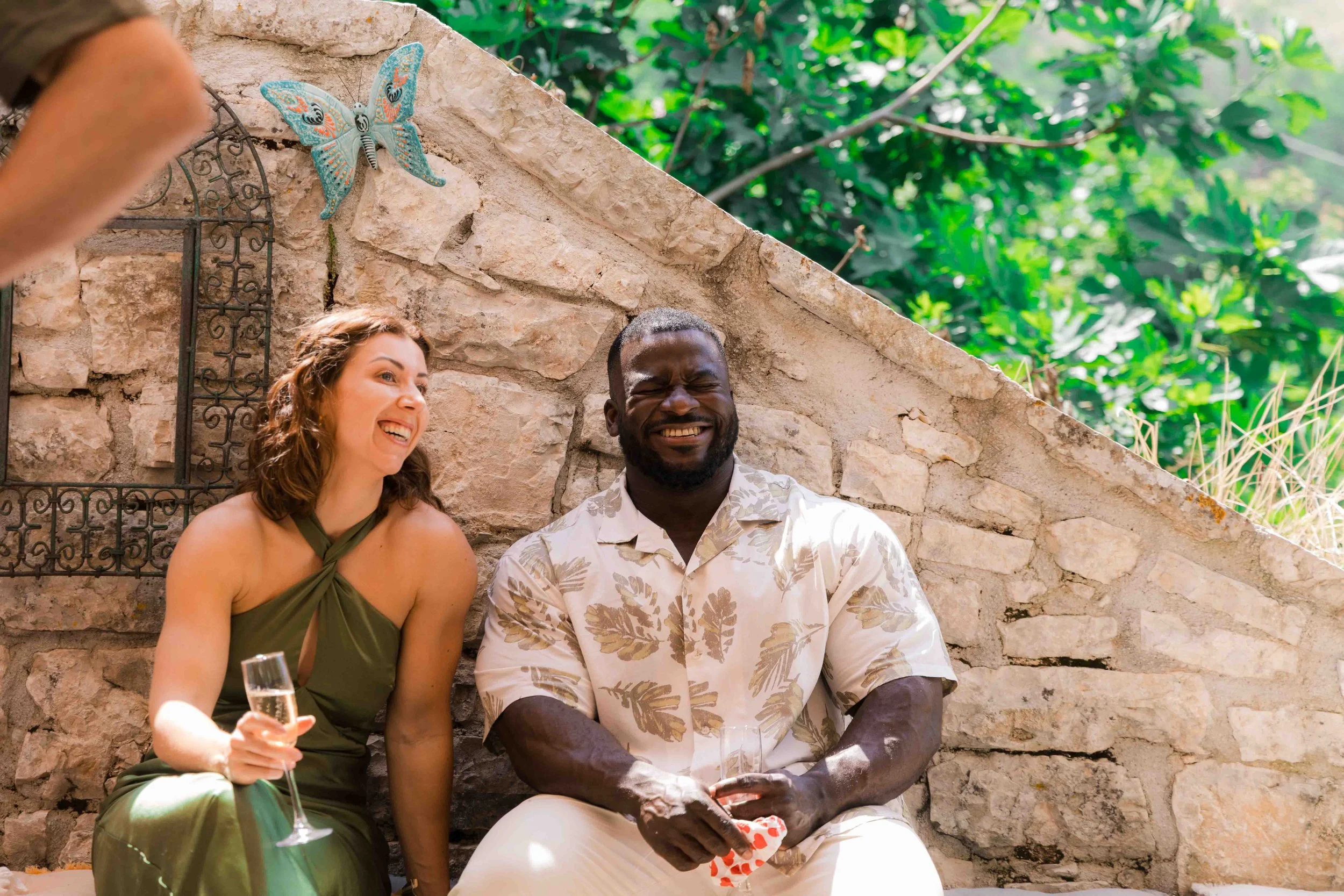 A woman and a man sitting outdoors against a stone wall, smiling and laughing, holding glasses of champagne, with green foliage behind them.