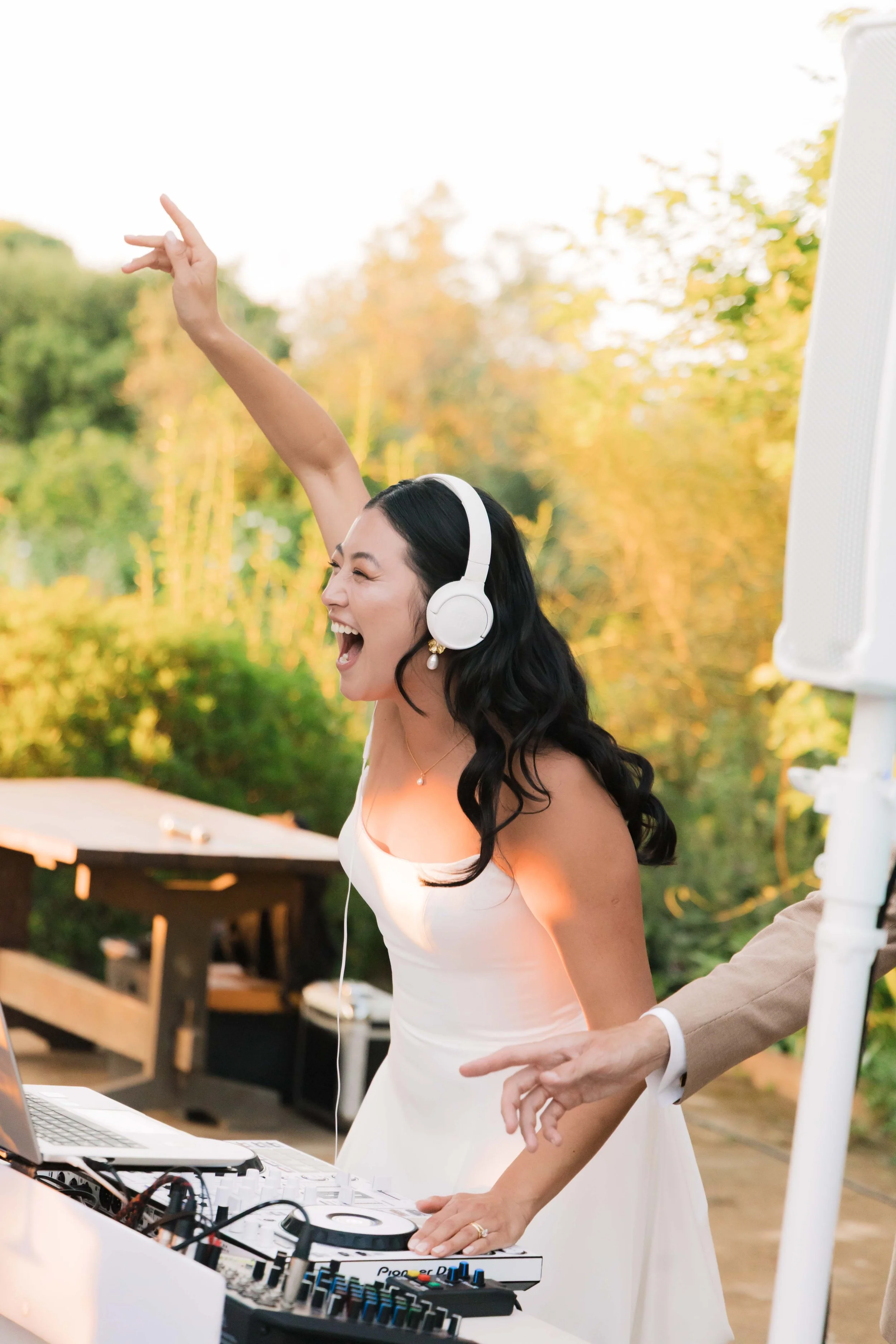 A woman in a white dress, wearing white headphones, dancing and having fun at an outdoor event with DJ equipment, green trees, and golden sunlight in the background.