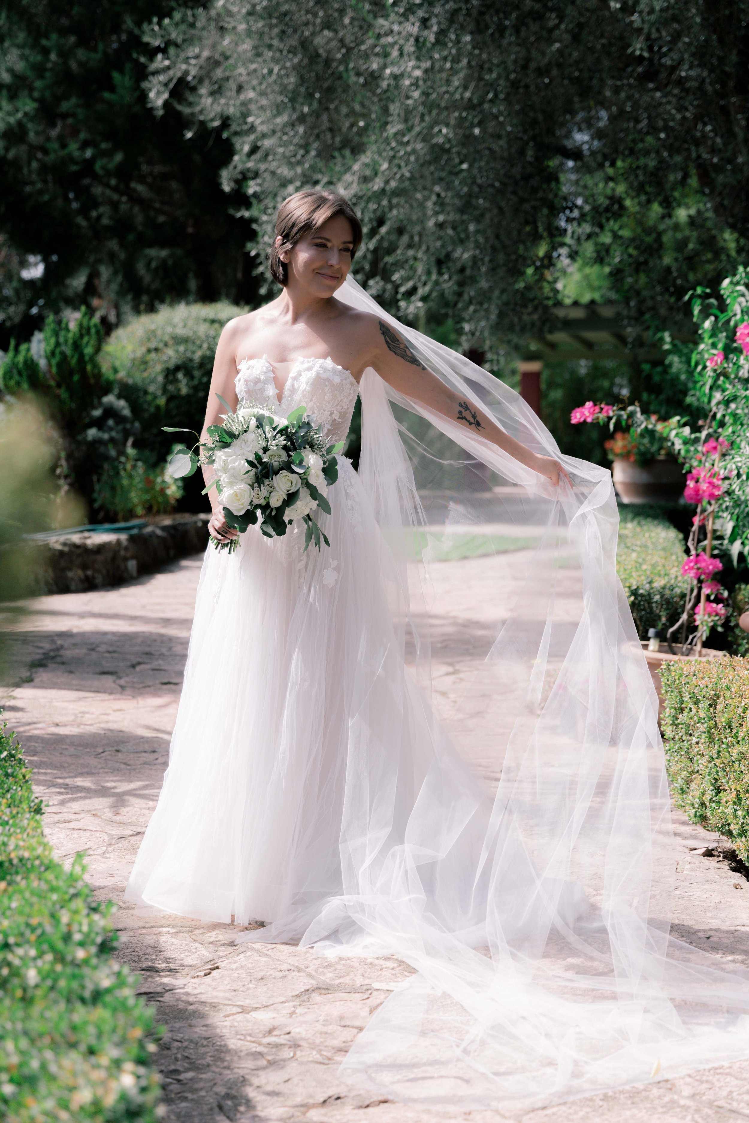 A bride in a white wedding dress holding a bouquet of white roses walking outdoors on a stone pathway, with greenery and pink flowers in the background.