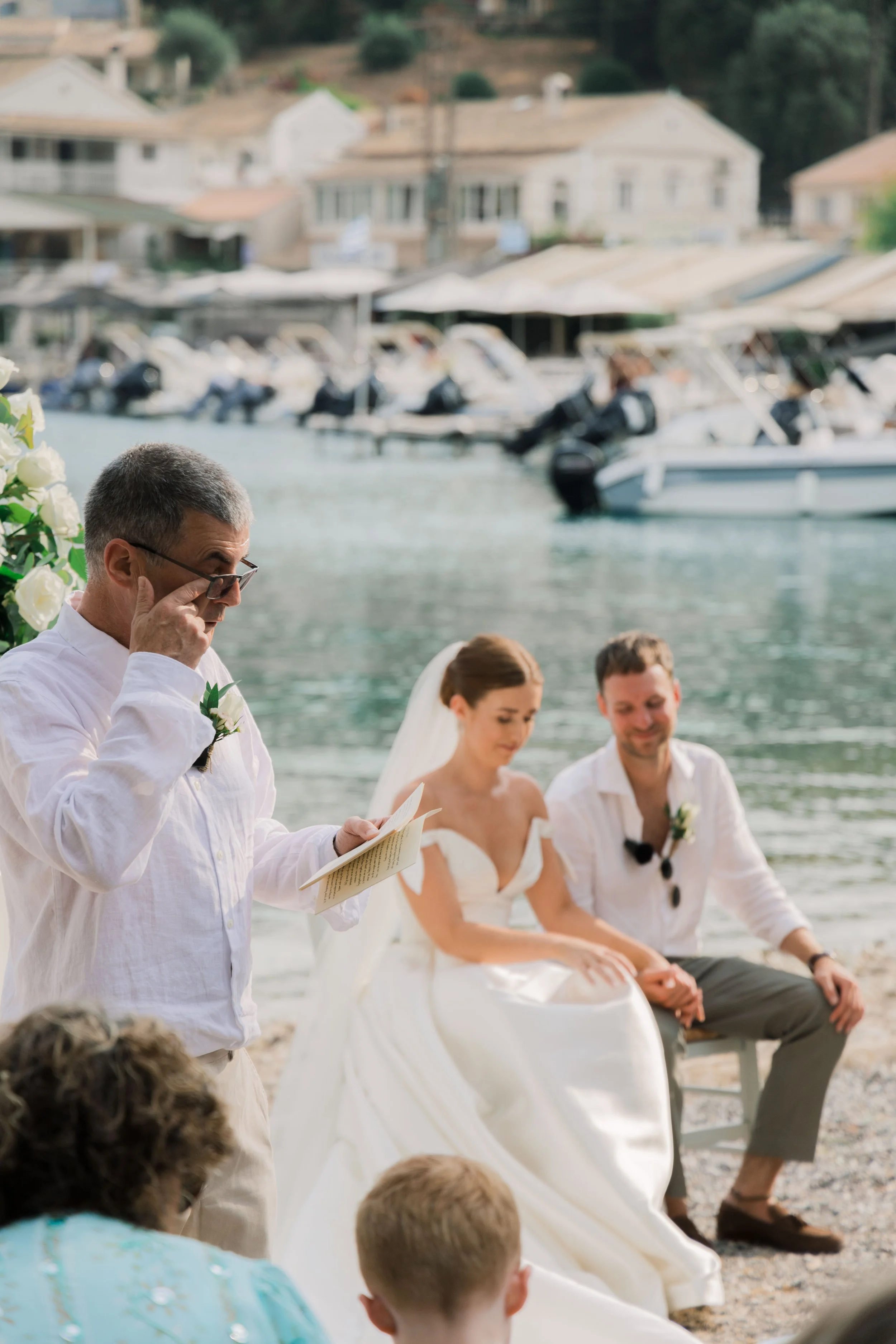 A wedding ceremony taking place outdoors on a lakeshore with boats and houses in the background. A man is reading from a paper, and a bride and groom sit nearby, holding hands and listening.