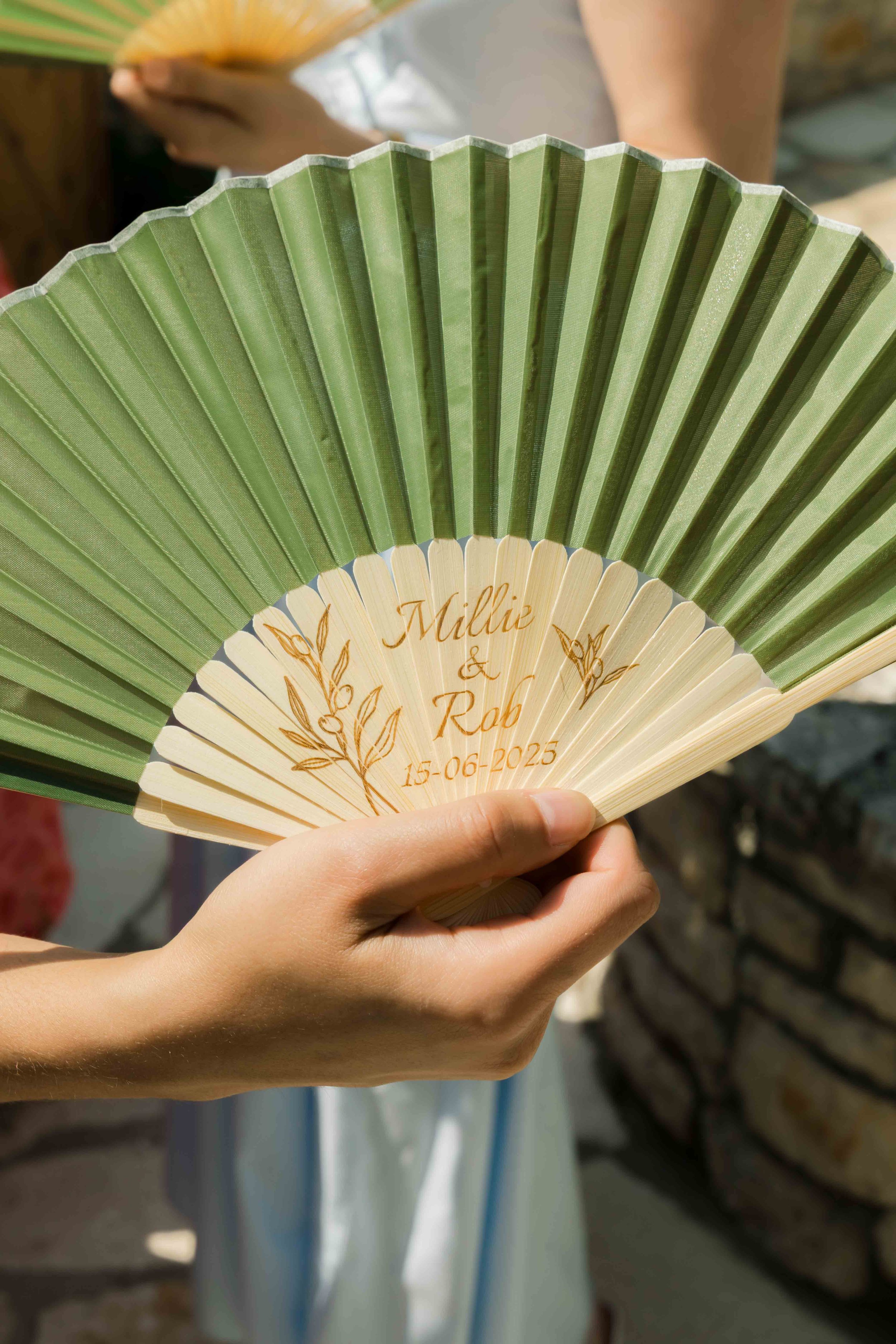 Close-up of a hand holding a decorative fan with green and beige sections, engraved with the names 'Millie & Rob' and the date '15-06-2025'.