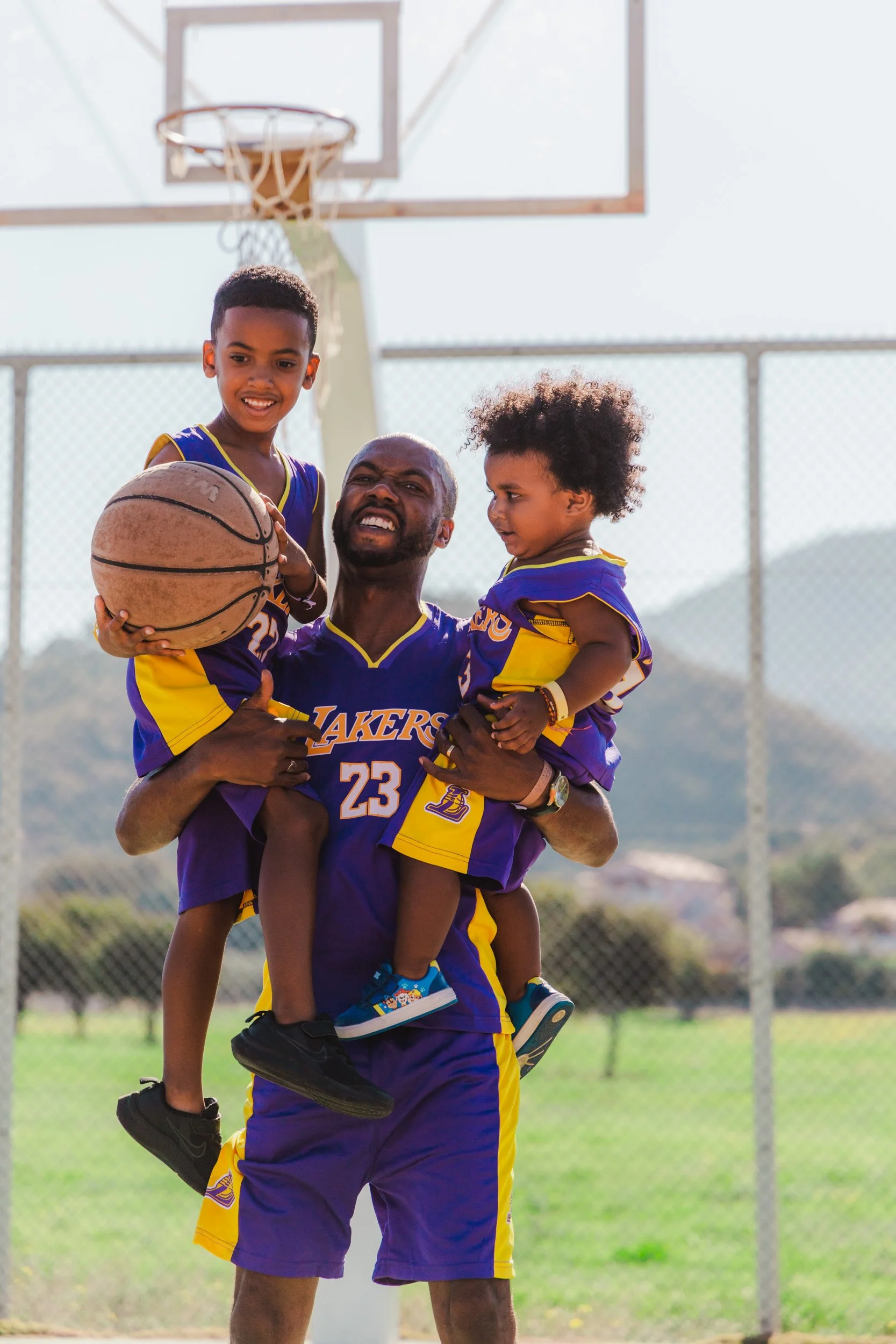 A man in a Los Angeles Lakers basketball uniform holding two children, all smiling, on a basketball court outdoors.