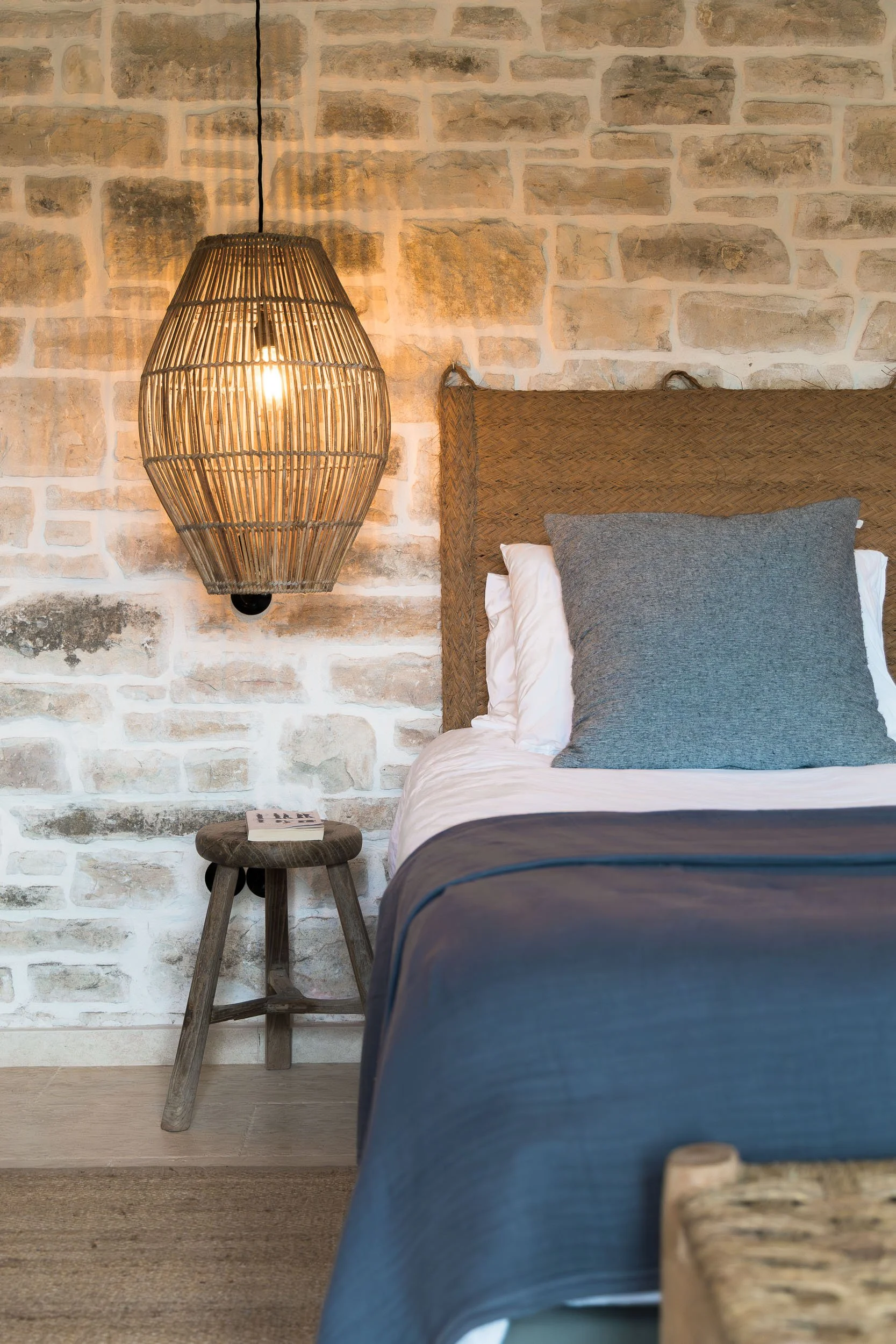 A cozy bedroom with a stone accent wall, a rattan headboard, a gray pillow, white sheets, a navy blue bedspread, a small wooden stool, and a woven pendant light hanging from the ceiling.