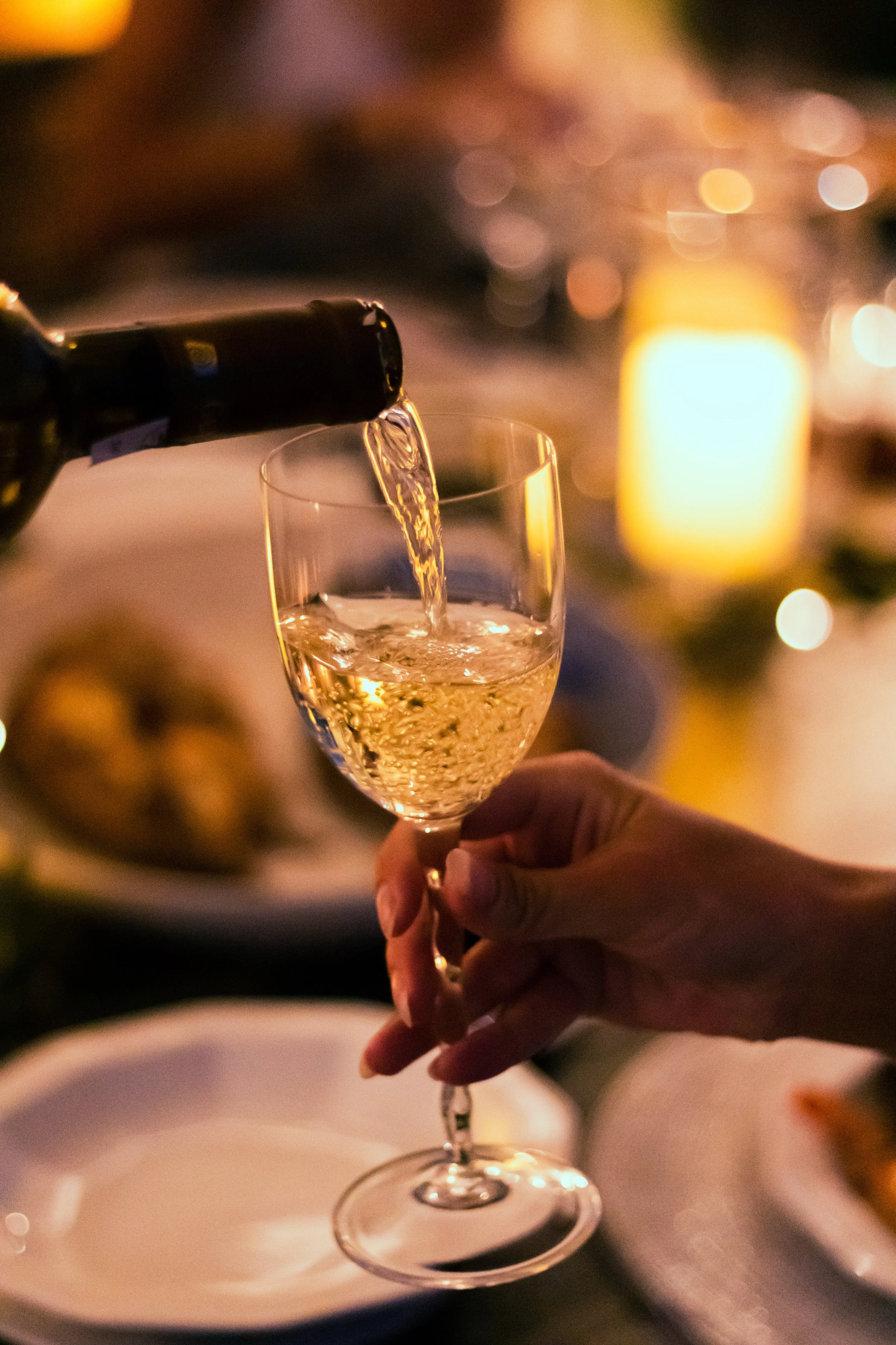 Person pouring white wine into a wine glass at a dinner table with blurred background