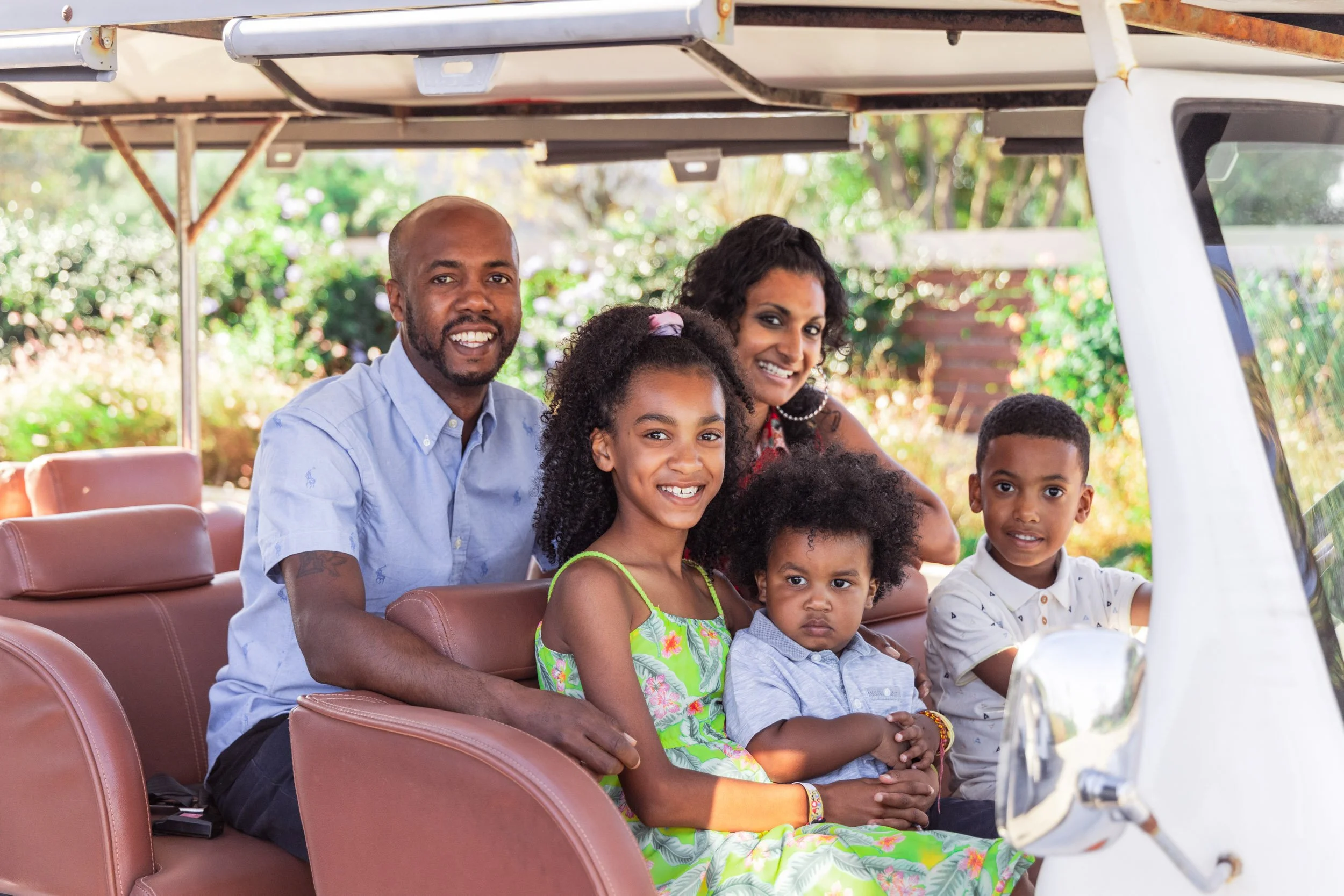 A family of six sitting together in a golf cart outdoors, smiling at the camera, with greenery in the background.