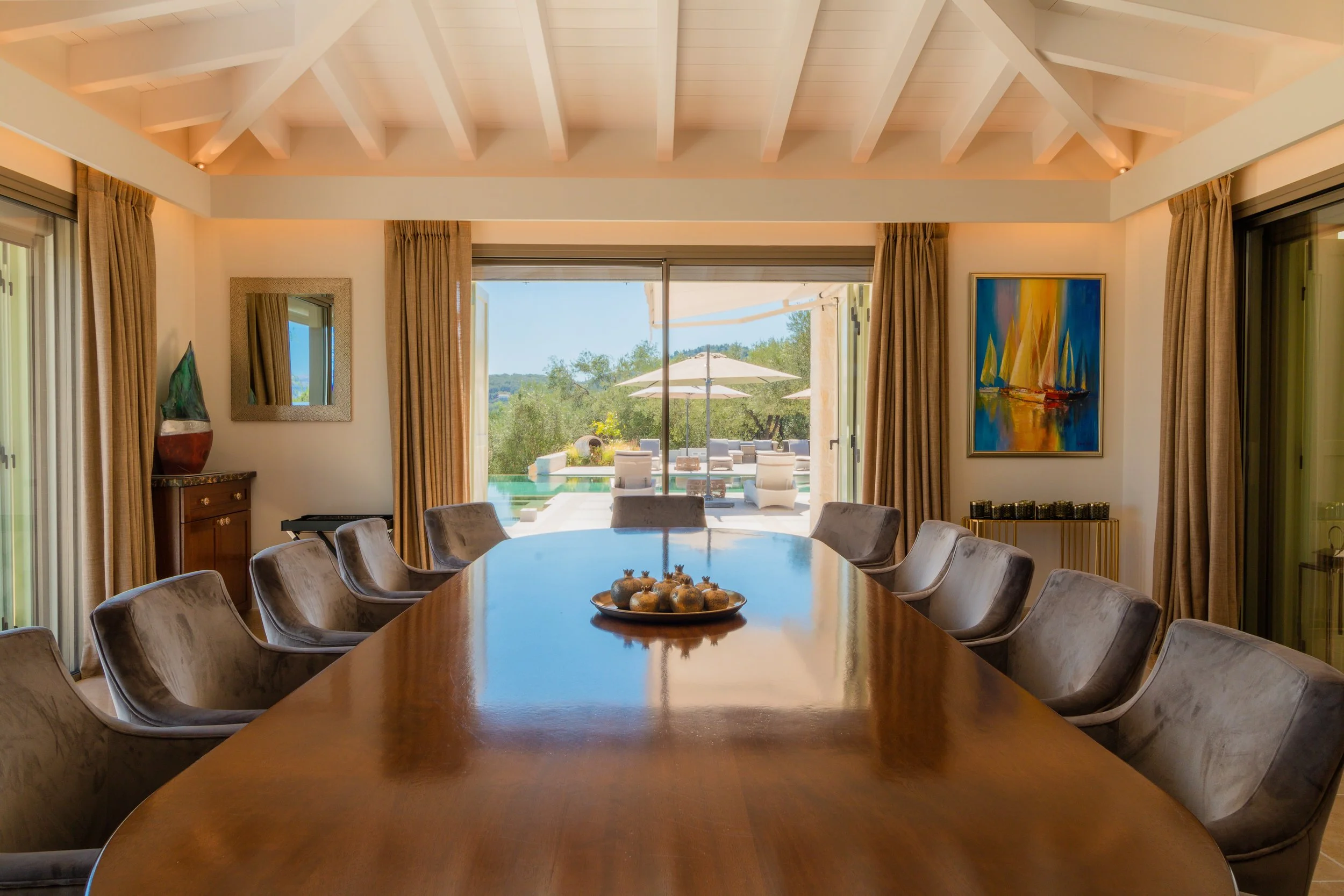 Luxury dining room with a long wooden table, velvet chairs, and a view of a patio and swimming pool through large sliding glass doors.