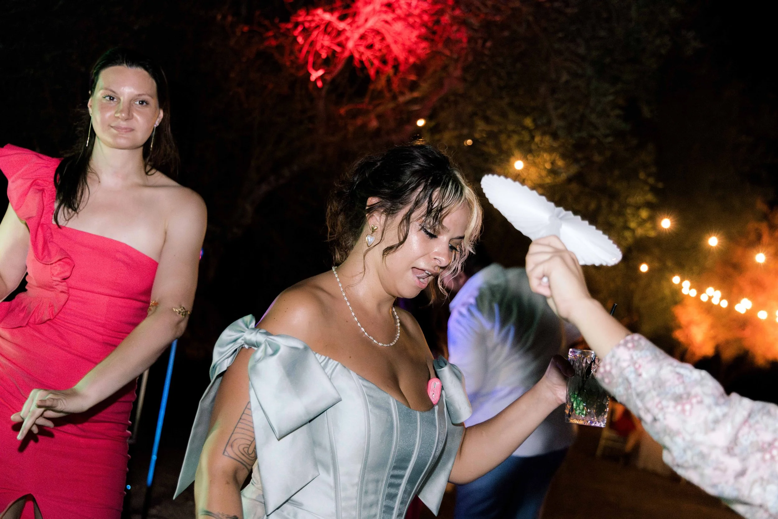At a nighttime outdoor party, two women are visible. One woman in a pink dress stands to the left, and another woman in a silver dress is in the center, holding a paper umbrella. The background features colorful lights and trees.