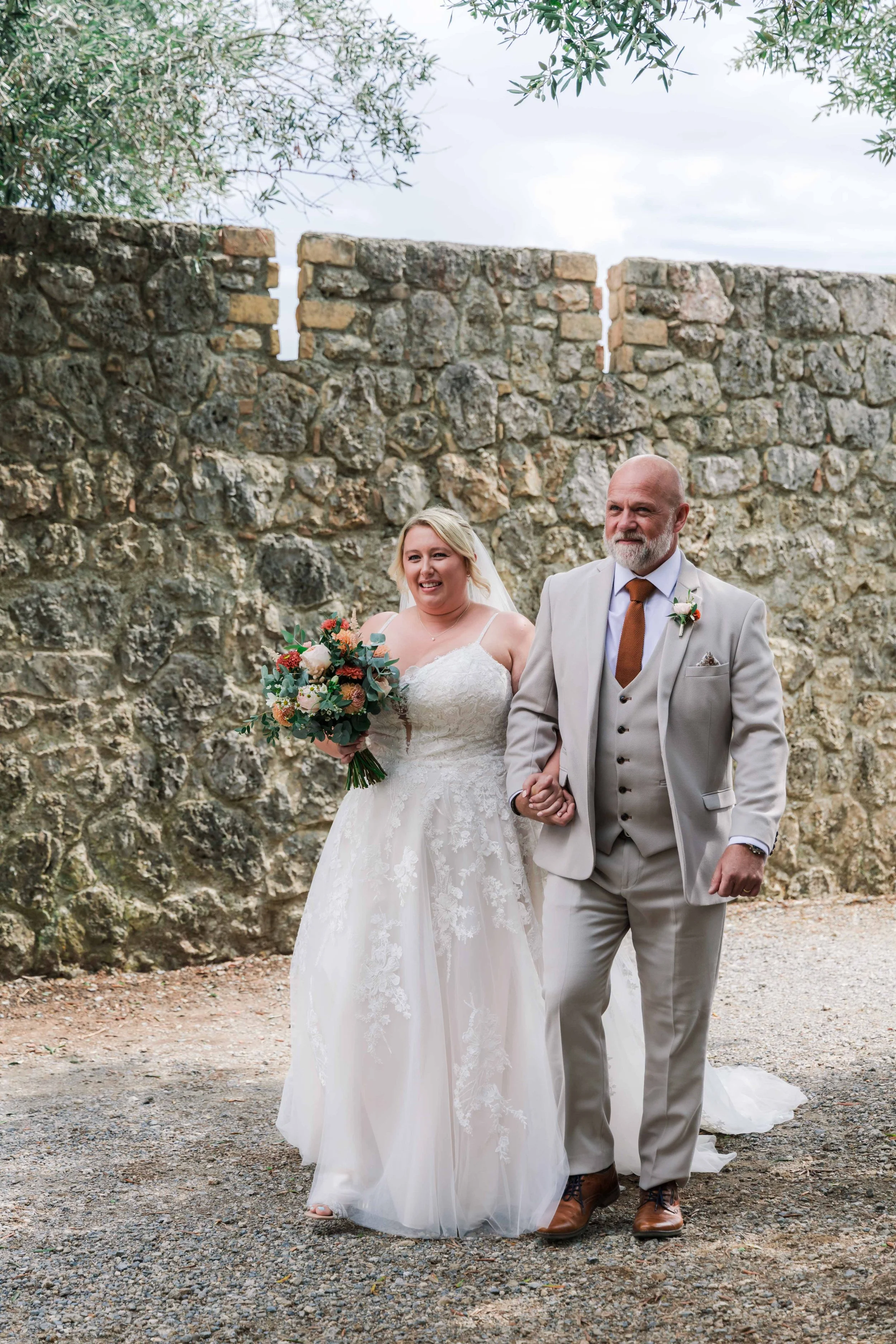 A bride in a white lace wedding dress holding a bouquet of flowers walking arm-in-arm with a man in a light gray suit and brown shoes, against a stone wall backdrop.