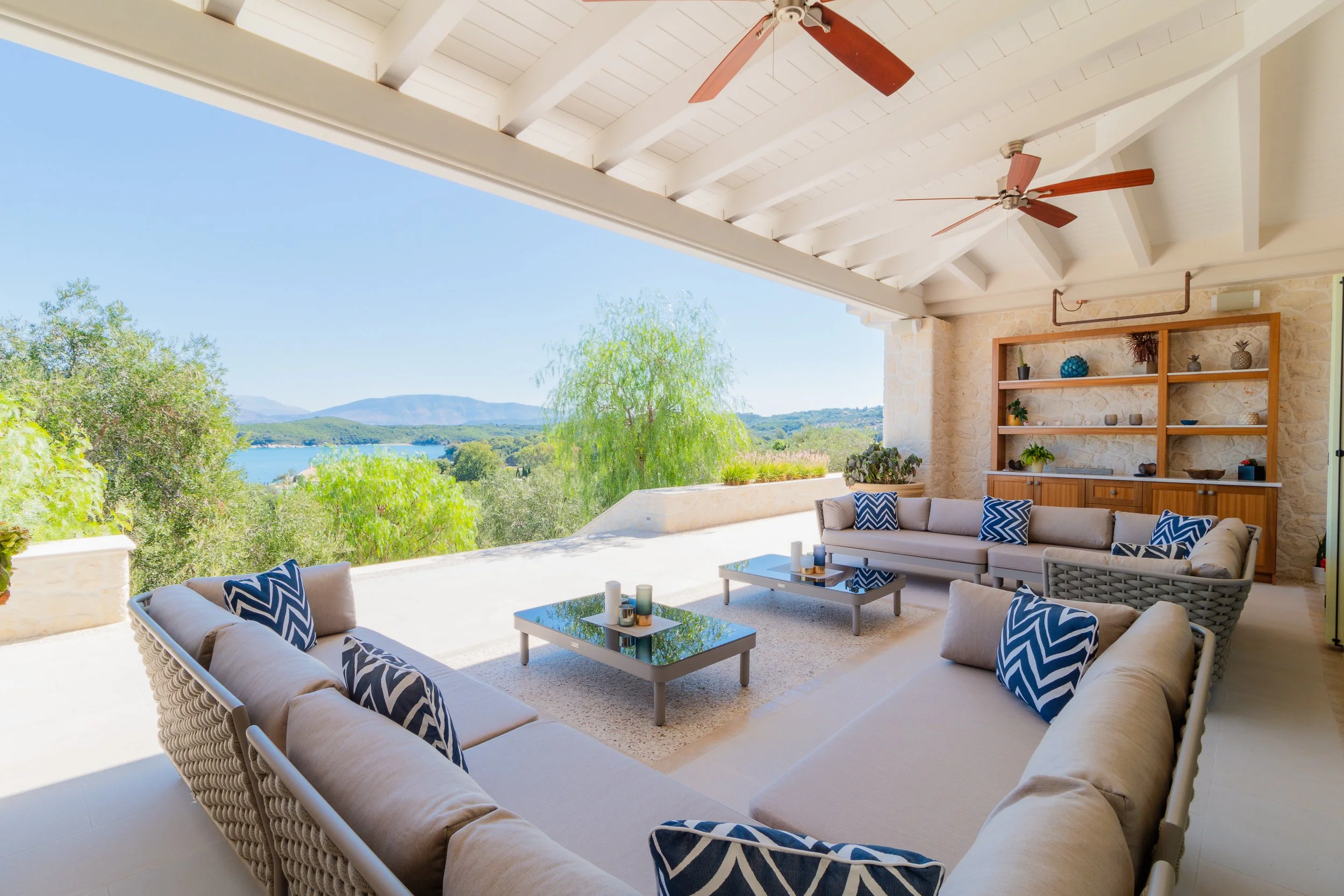 Outdoor living space with beige sectional sofas, blue and white chevron pillows, two coffee tables with candles, overlooking a scenic landscape with trees and water, covered by a white ceiling with wooden beams and ceiling fans.