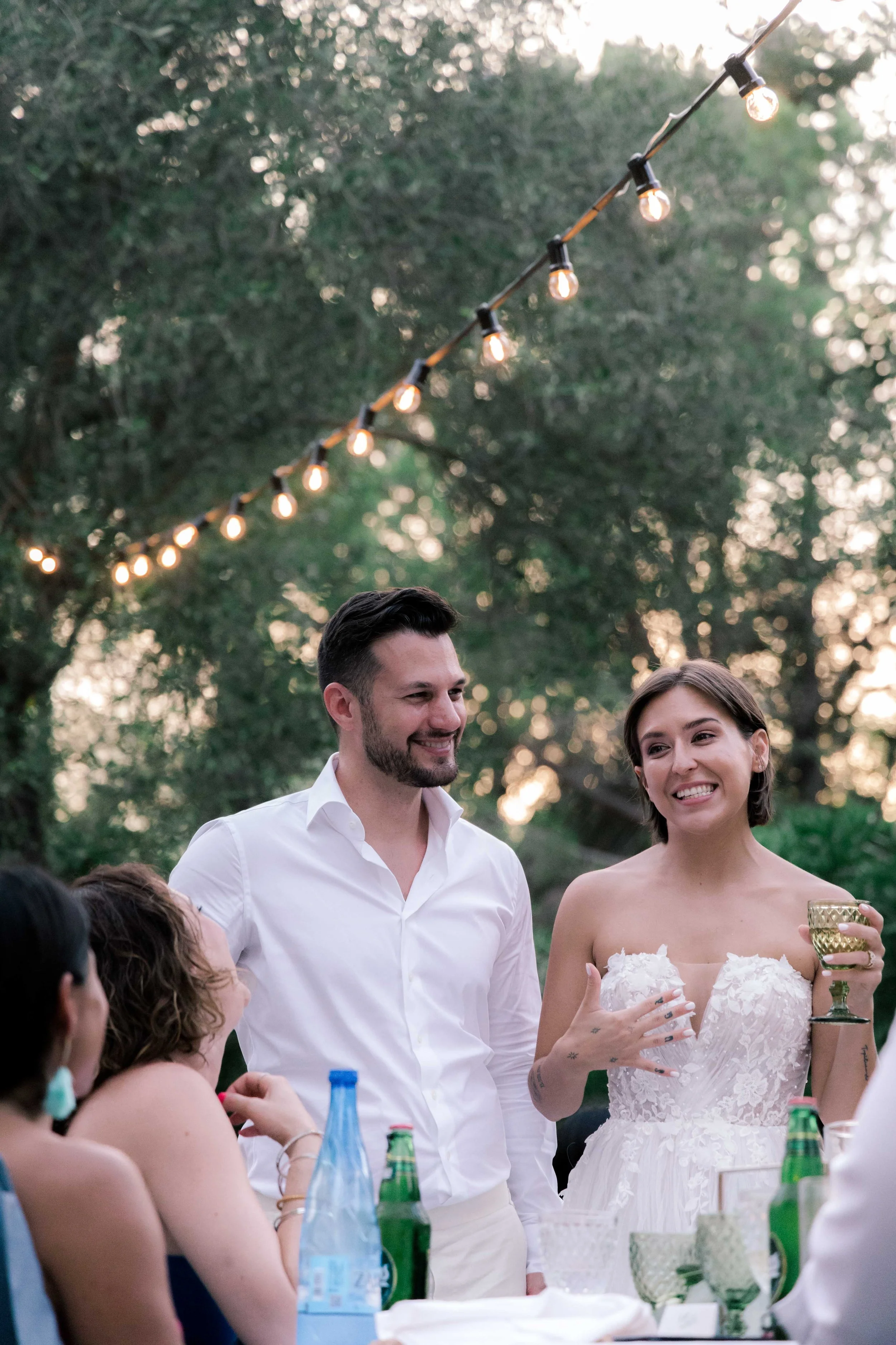A wedding reception outdoors with guests seated at tables, string lights hanging above, and a smiling bride holding a glass, while a groom stands beside her.