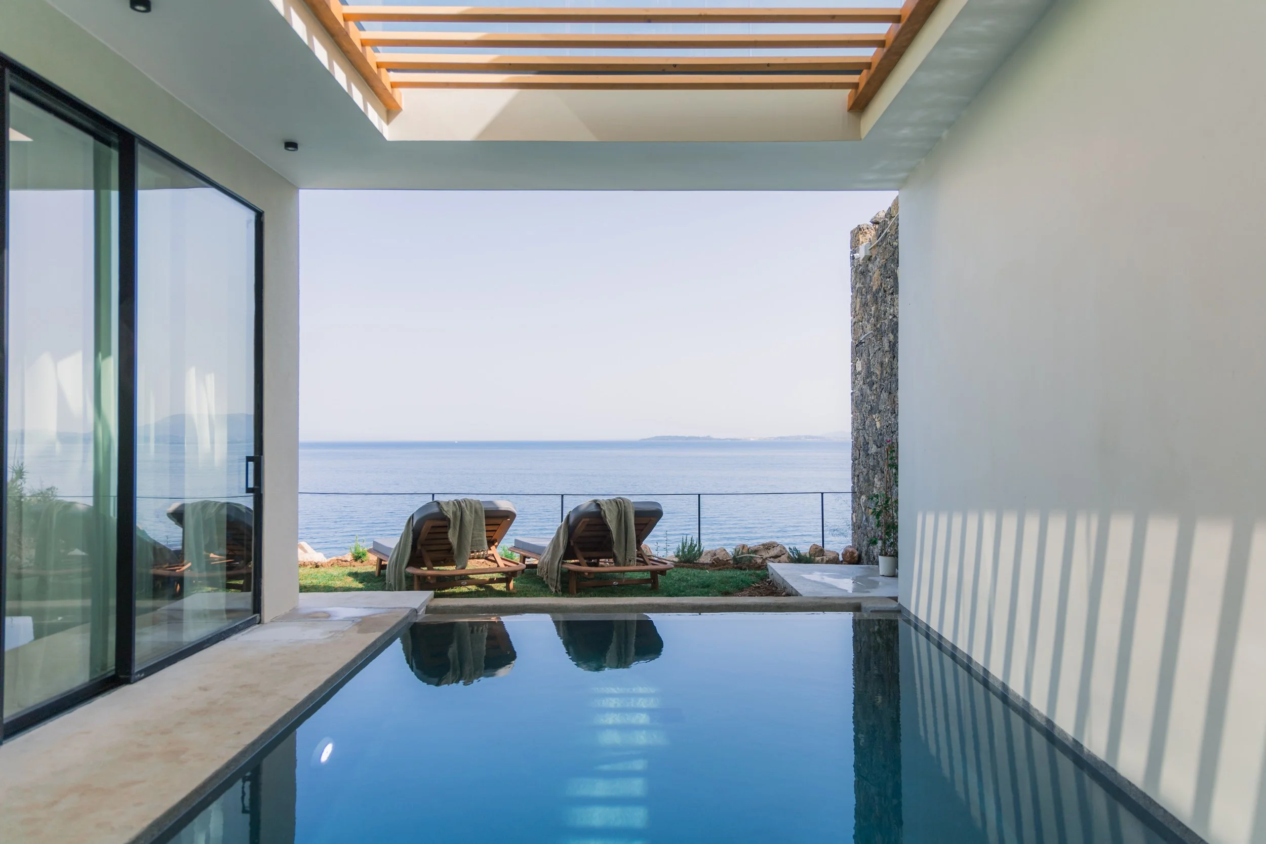 Indoor pool area with large glass sliding doors opening to a view of the ocean and two lounge chairs outside, overlooking the water.