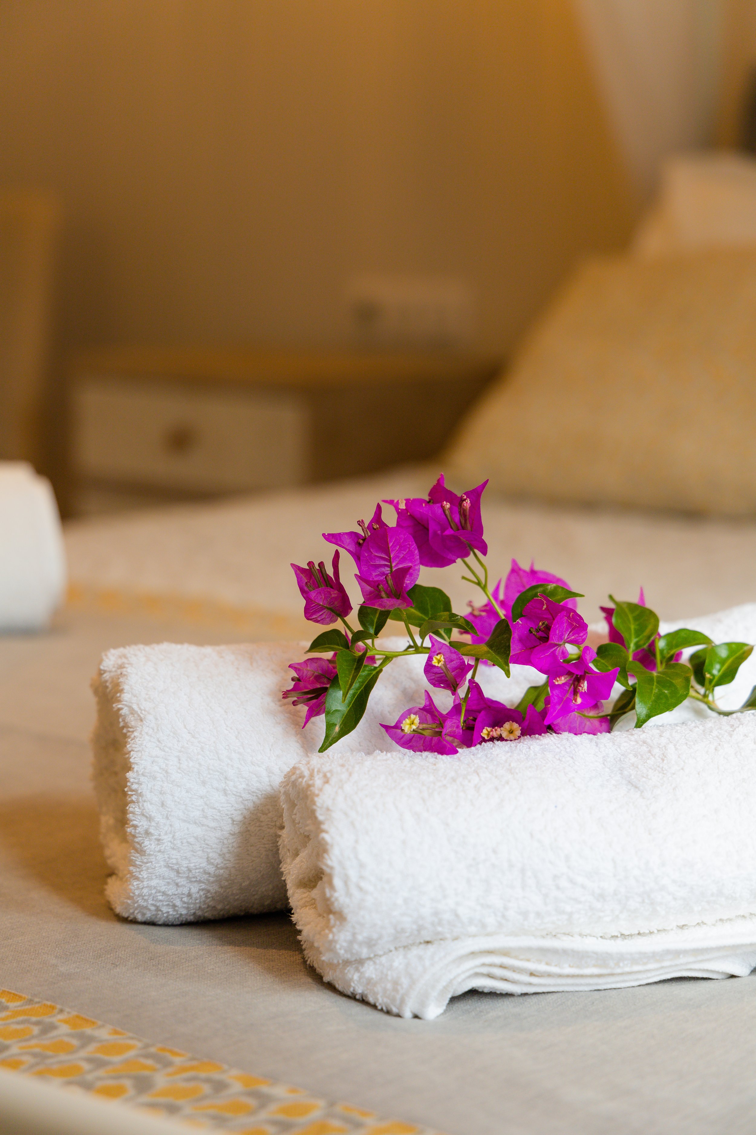 Decorative arrangement of pink bougainvillea flowers on a rolled white towel on a bed in a warmly lit room.