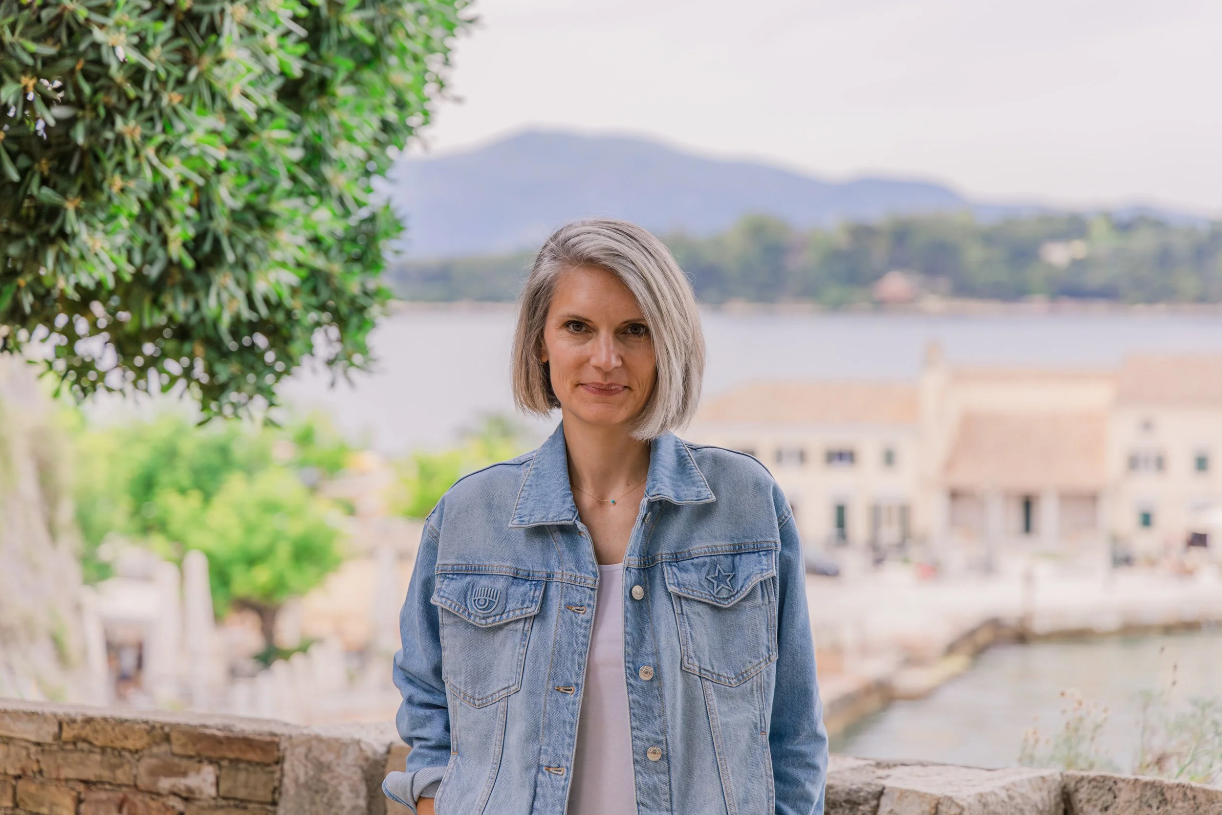 A woman with short gray hair standing outdoors near water with houses and mountains in the background, wearing a denim jacket.