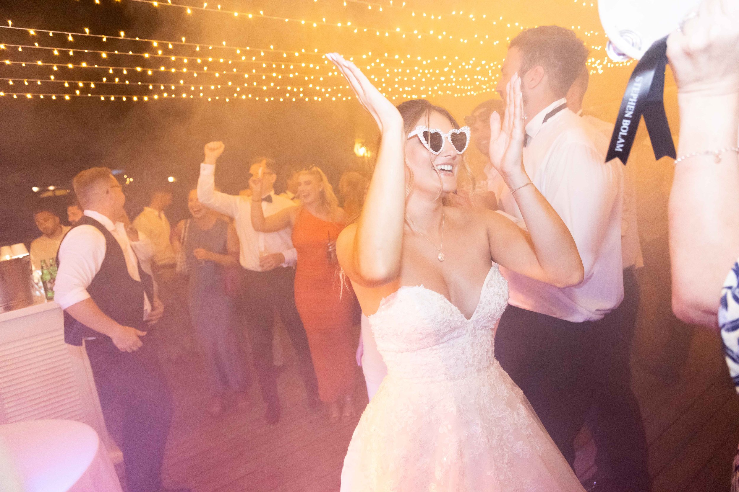 Bride dancing with guest at a wedding reception, surrounded by guests holding drinks and celebrating under string lights.