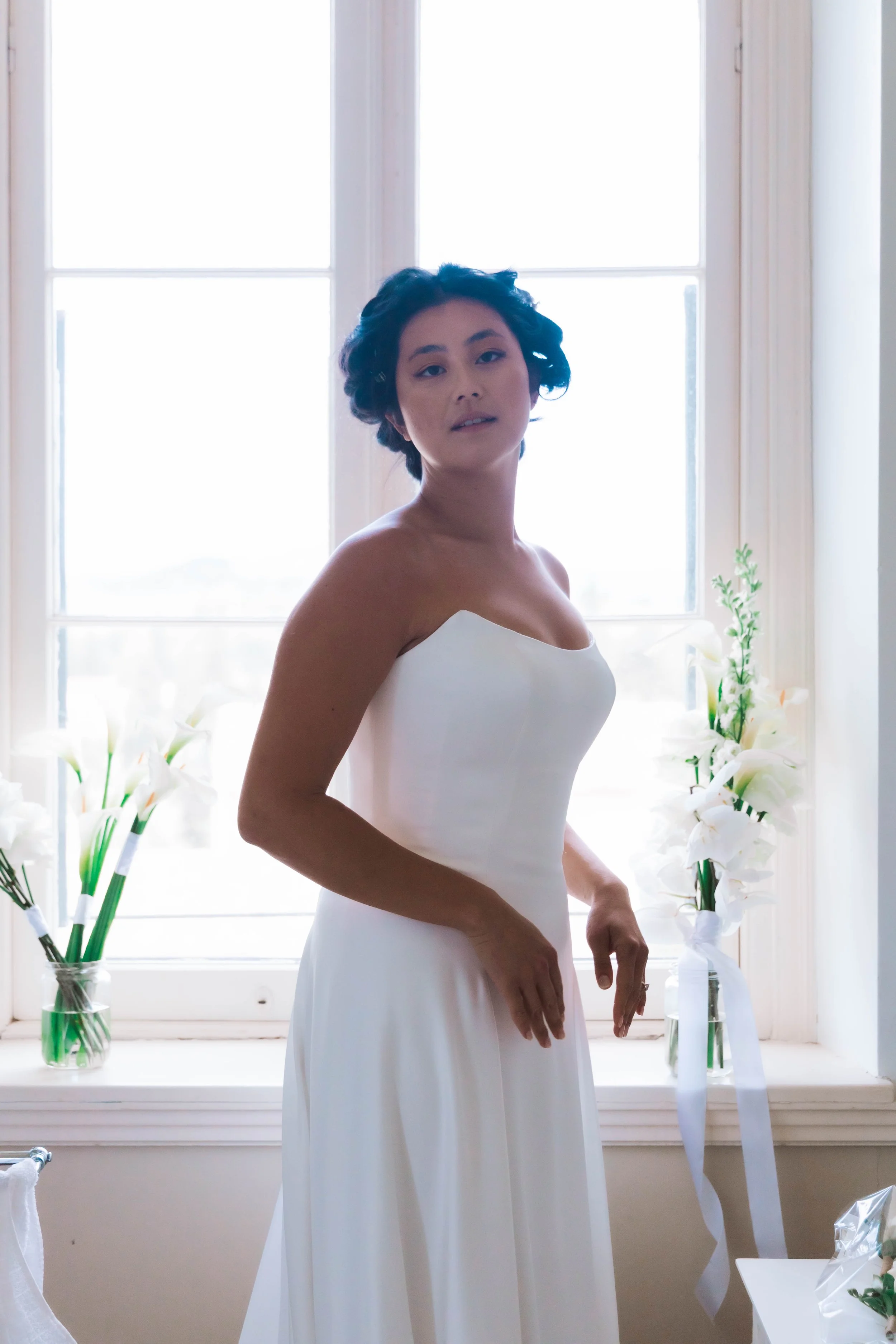 Woman in a strapless white gown standing by a window with white flowers on the sill.