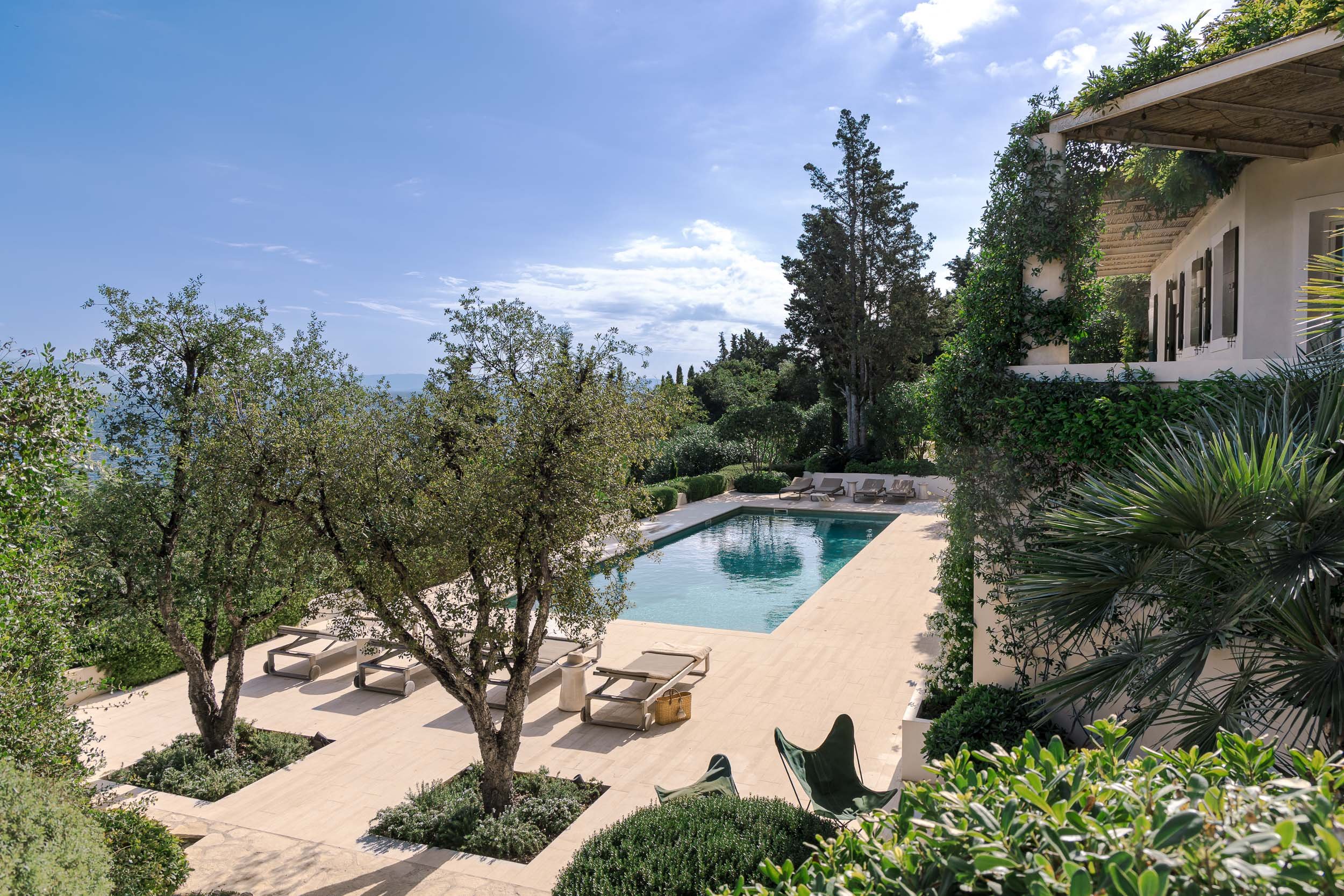 Outdoor swimming pool surrounded by lounge chairs, trees, and lush greenery, with a white building on the right and a clear blue sky overhead.