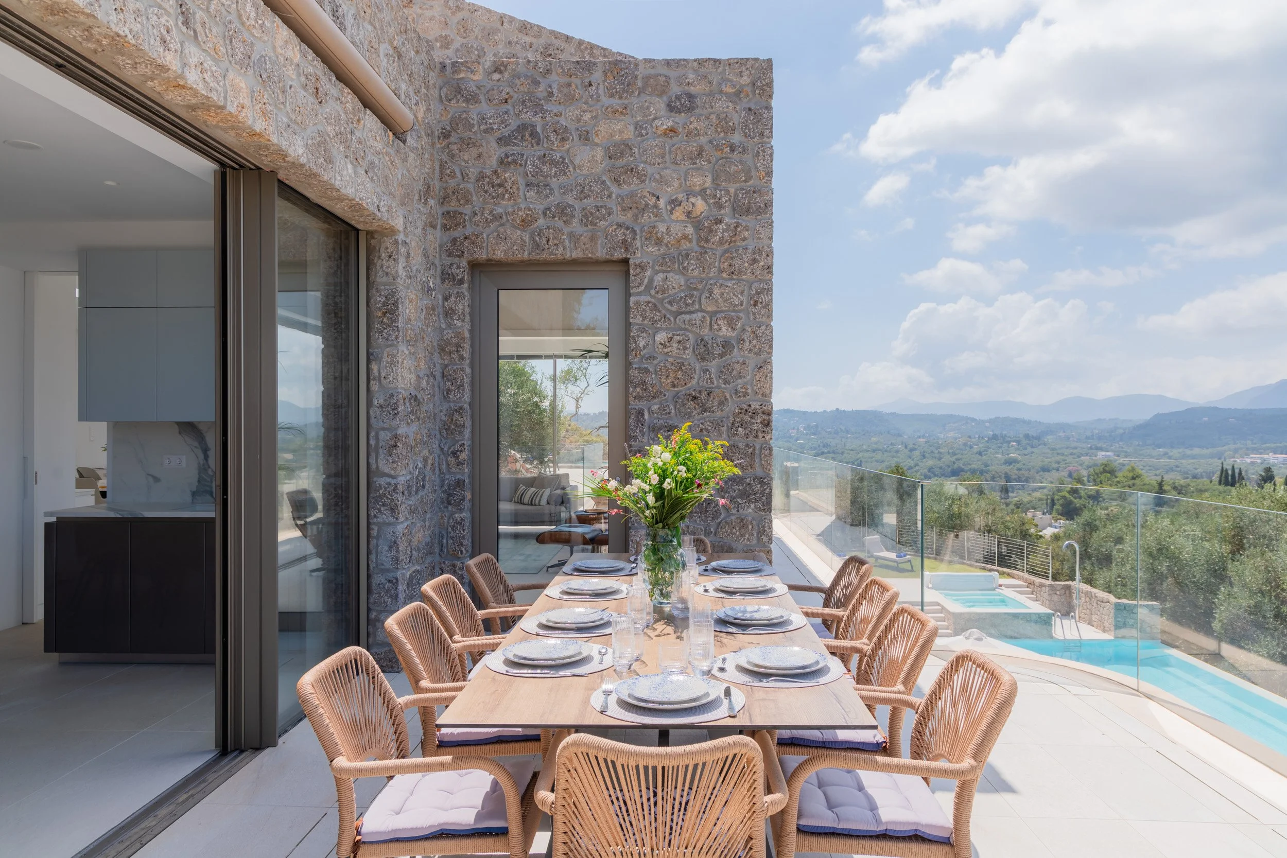 Outdoor dining table set for eight with a vase of flowers, overlooking a pool and scenic landscape with mountains.