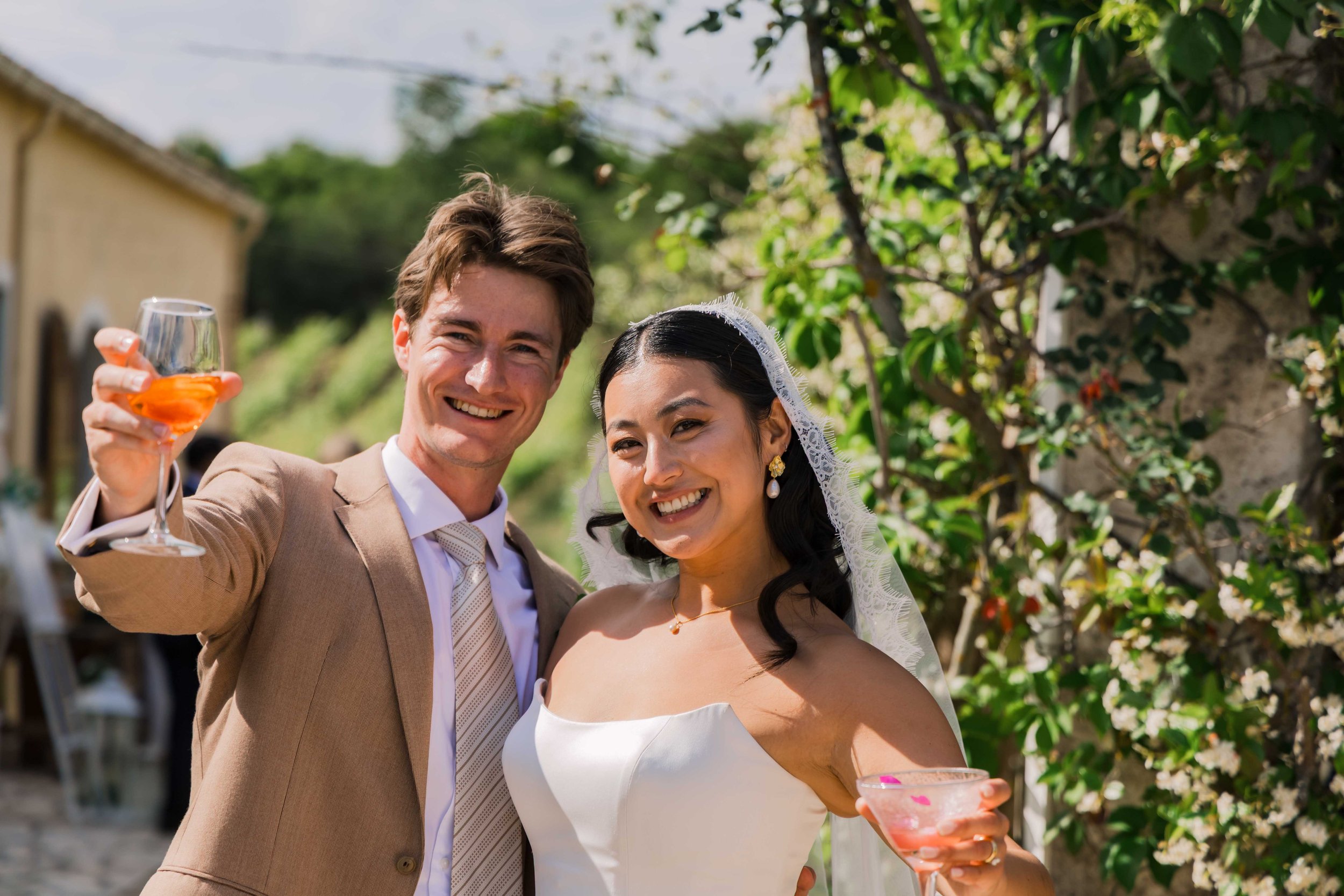 A happy couple at a wedding celebration outdoors, holding glasses of wine or champagne. The groom is wearing a beige suit, and the bride is in a white strapless wedding dress with a veil, surrounded by greenery.