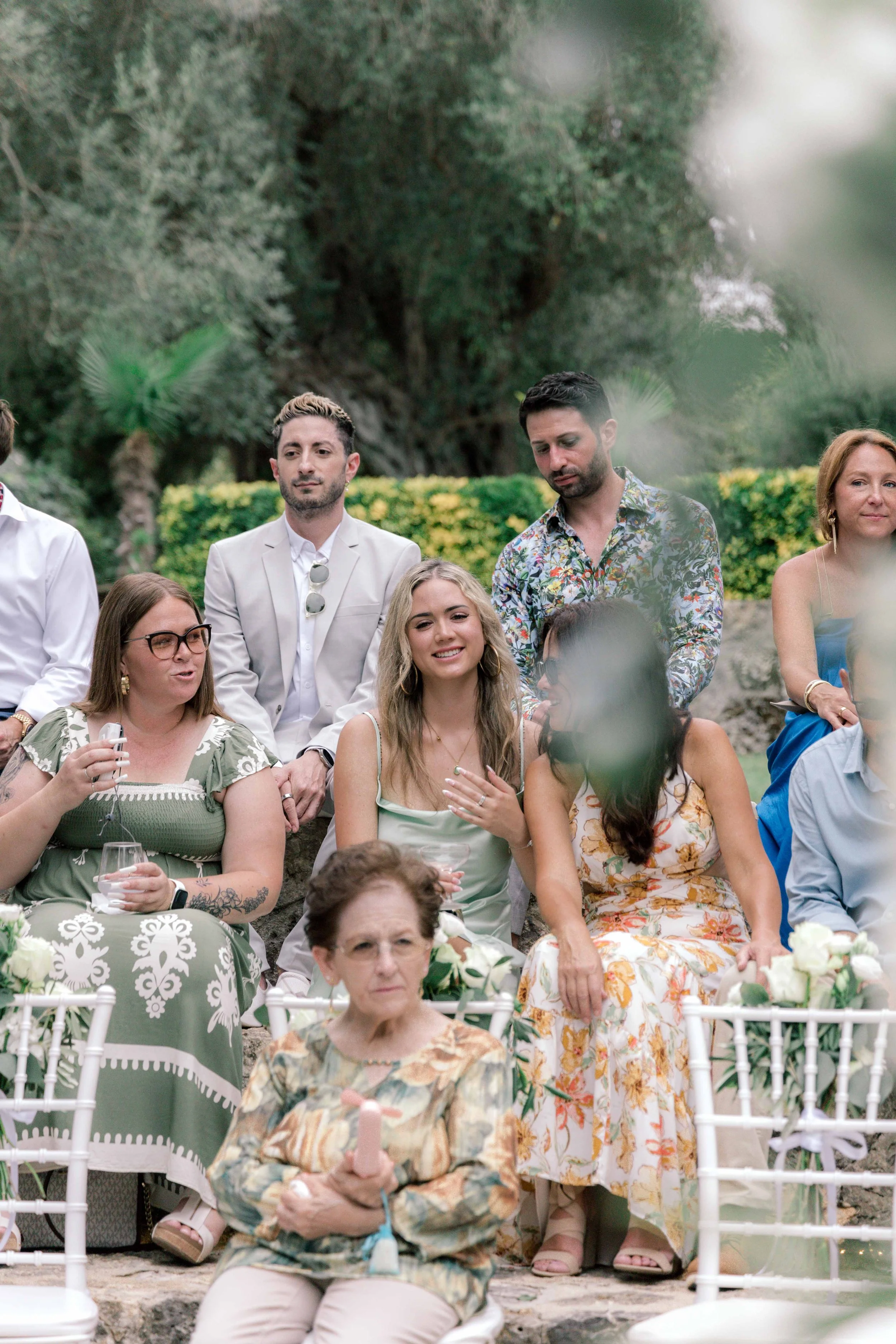 Group of people seated outdoors during a celebration, with greenery and trees in the background.