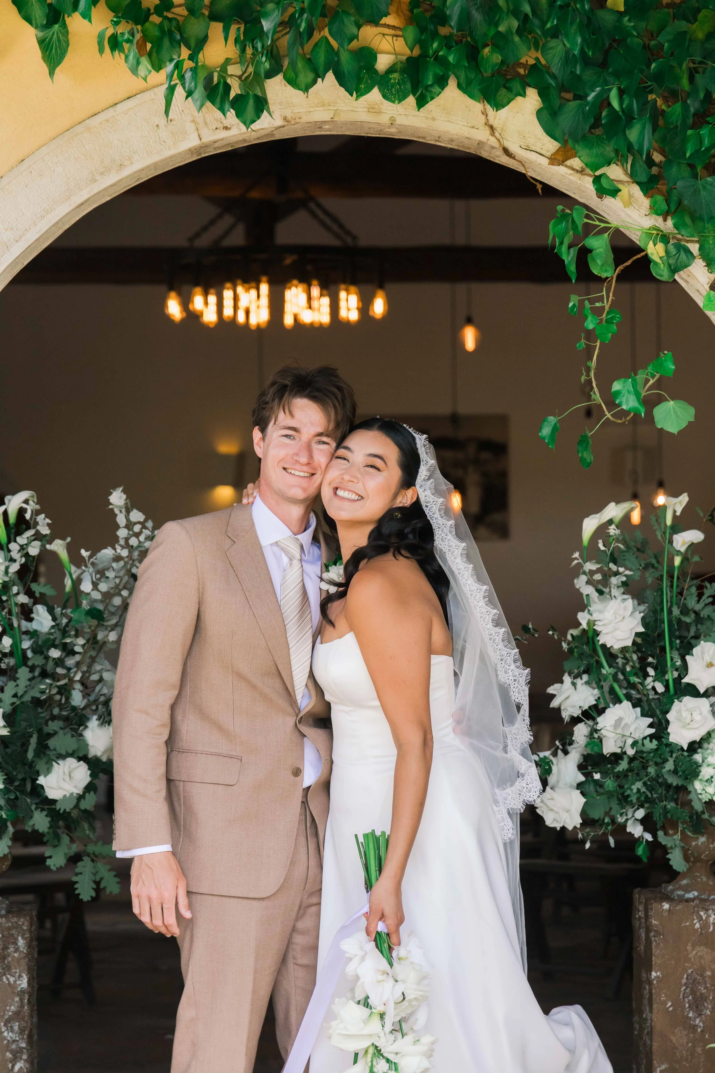 A newlywed couple in wedding attire, smiling and embracing each other, standing under an archway decorated with green leaves and white flowers, with warm lighting hanging above indoors.