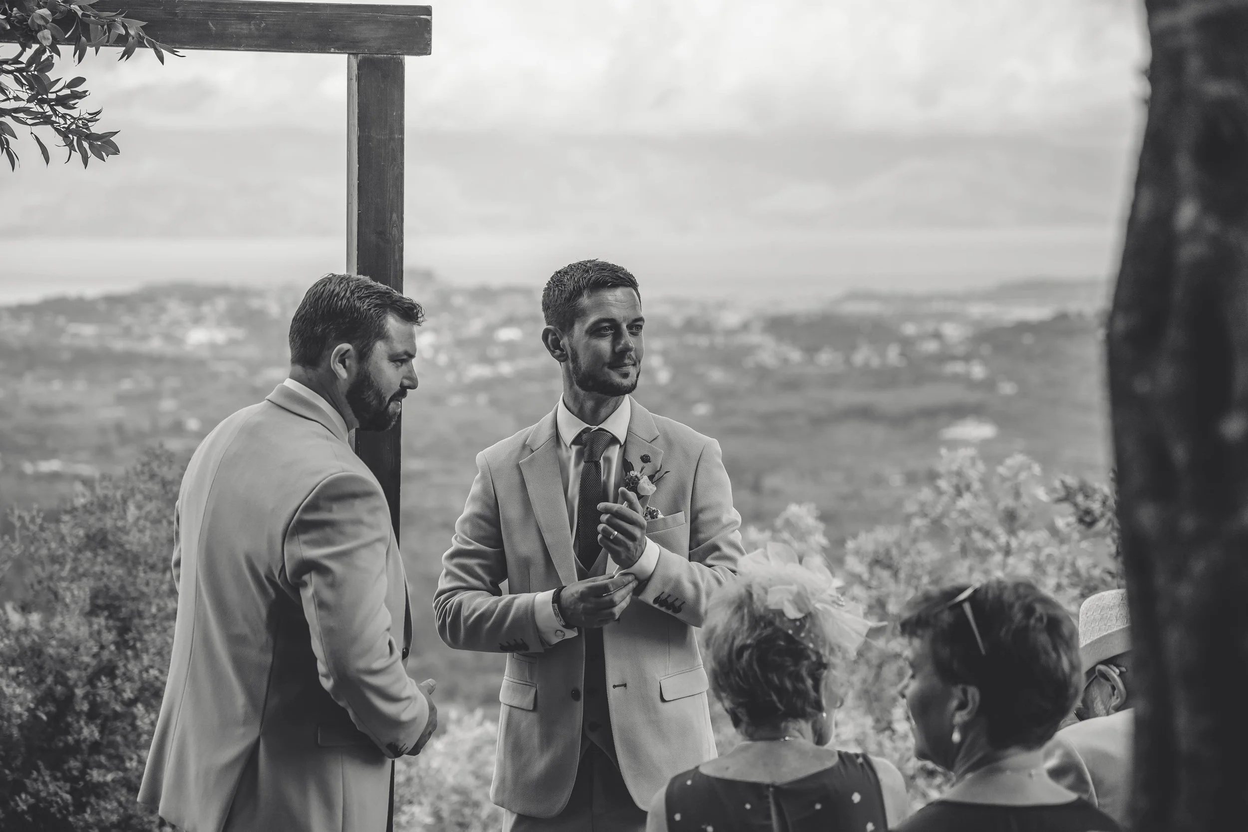 Black and white image of two men at a wedding ceremony, one of them is holding a small object, with guests seated in front and a scenic landscape in the background.