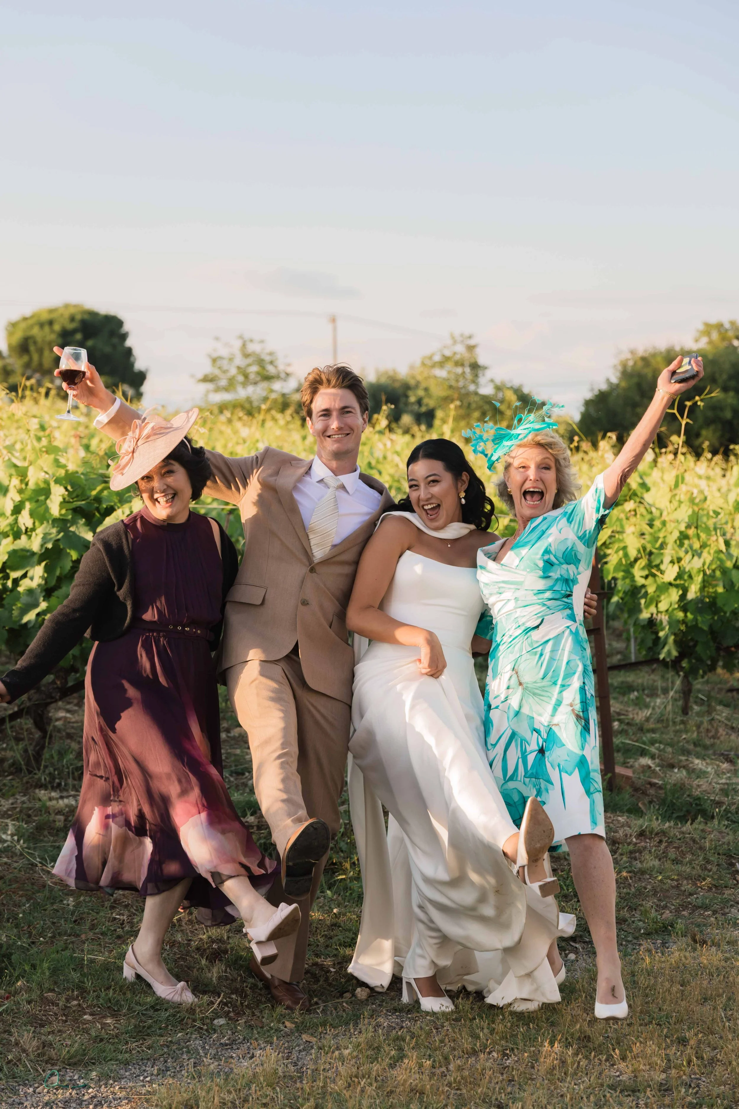 Group of five people, including a bride in a white wedding dress, celebrating outdoors in a vineyard, holding glasses of wine, with smiles and cheerful expressions.