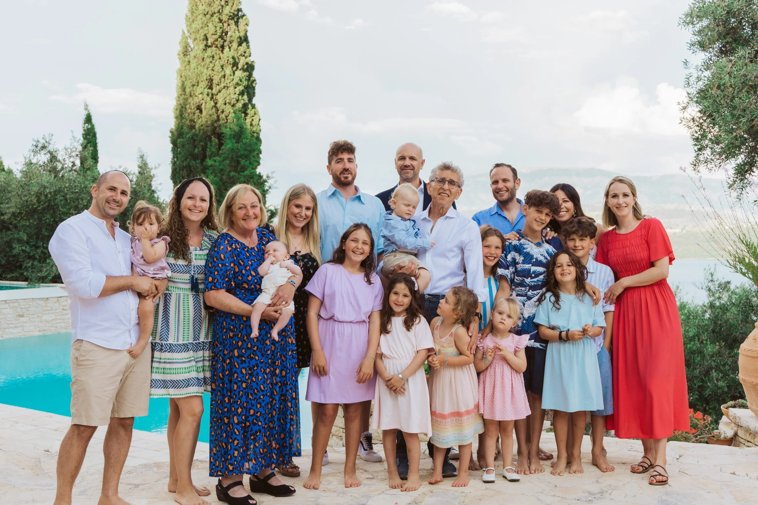 A large multi-generational family gathered outdoors by a swimming pool under a partly cloudy sky, smiling for a group photo.