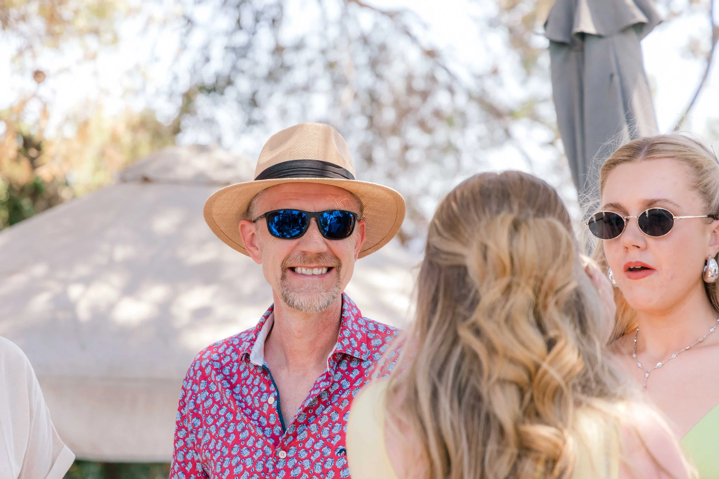 Smiling man wearing a straw hat and blue sunglasses standing outdoors, with two women nearby, one with blonde hair and sunglasses, in a sunny outdoor setting with trees in the background.
