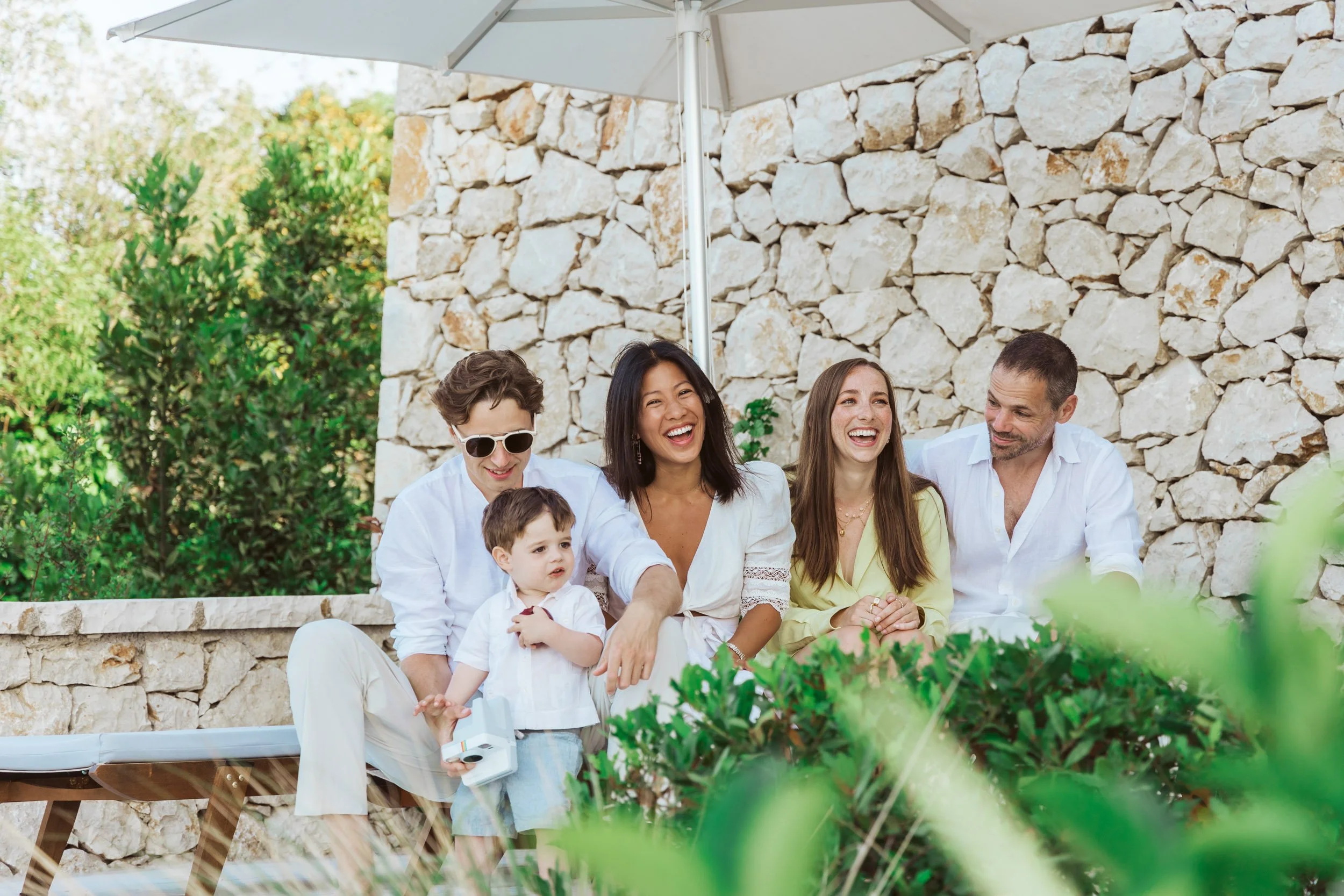 Group of five people sitting outdoors under a large white umbrella, smiling and enjoying each other's company, with a stone wall and greenery in the background.