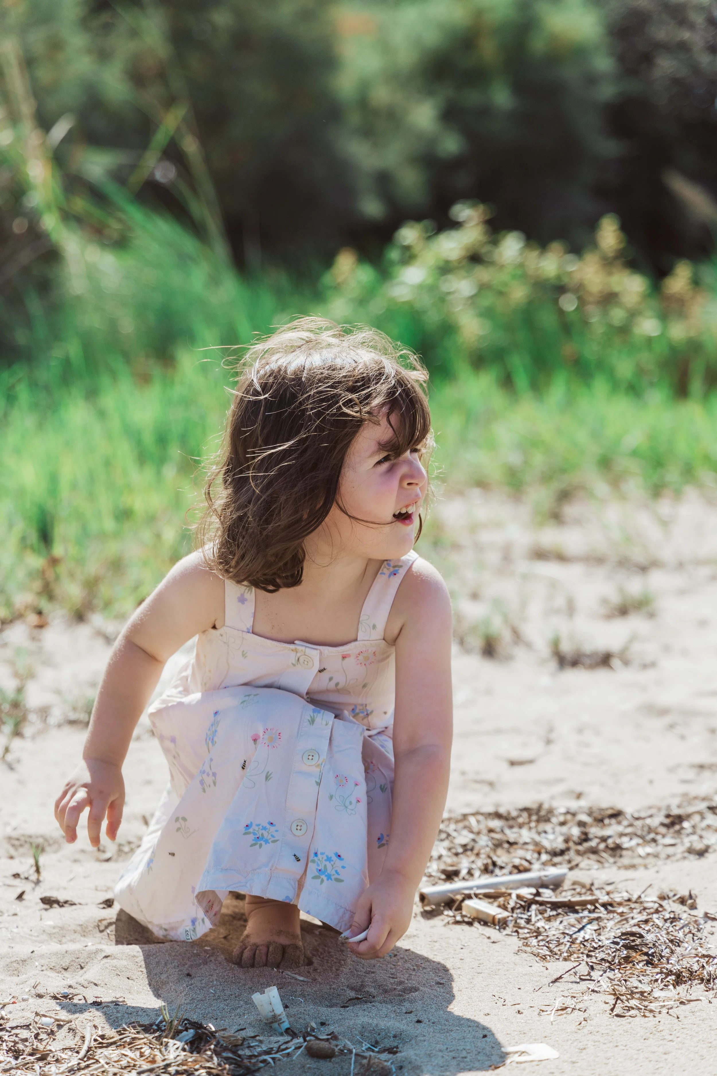 A young girl wearing a light-colored dress with floral patterns crouching on sandy ground outdoors in a natural setting with greenery in the background.