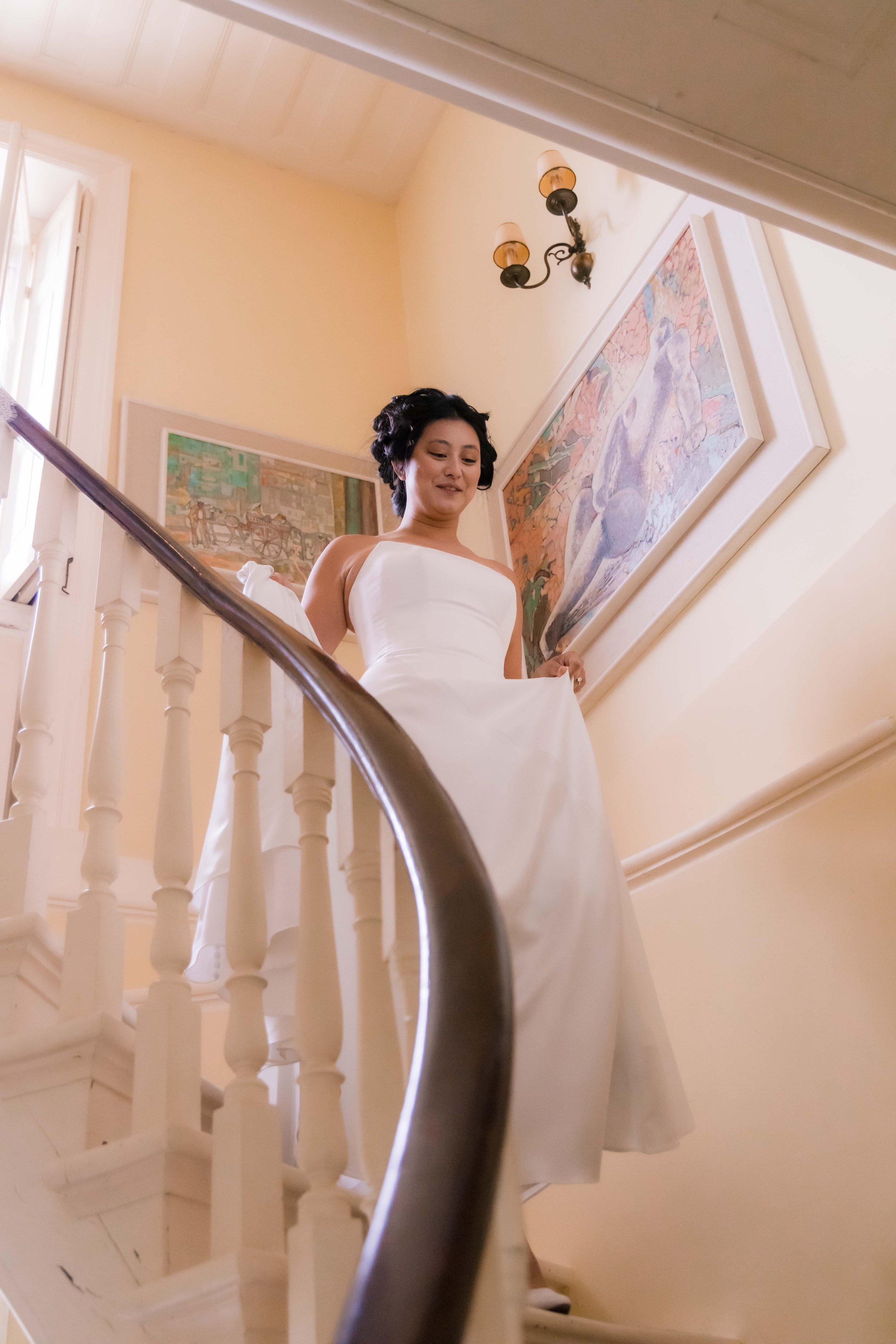 A woman in a white wedding dress standing on a staircase, looking down, with framed artwork on the wall behind her.