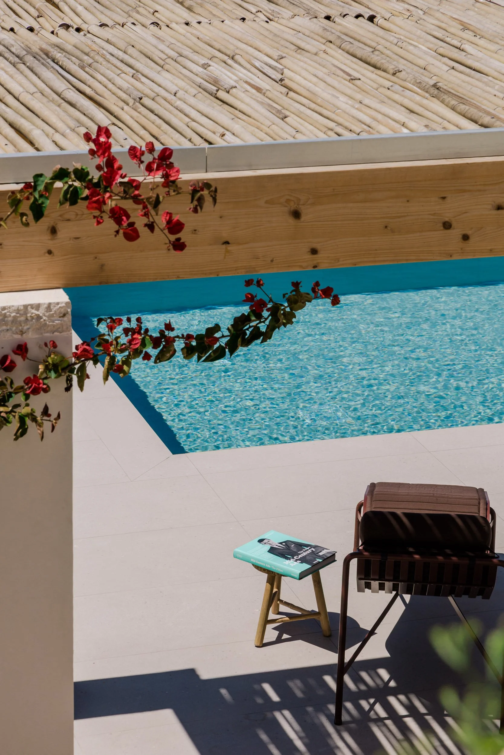 A backyard pool area with a swimming pool, a lounge chair with a book, and a small wooden stool. There are red flowering plants overhanging from the left side.
