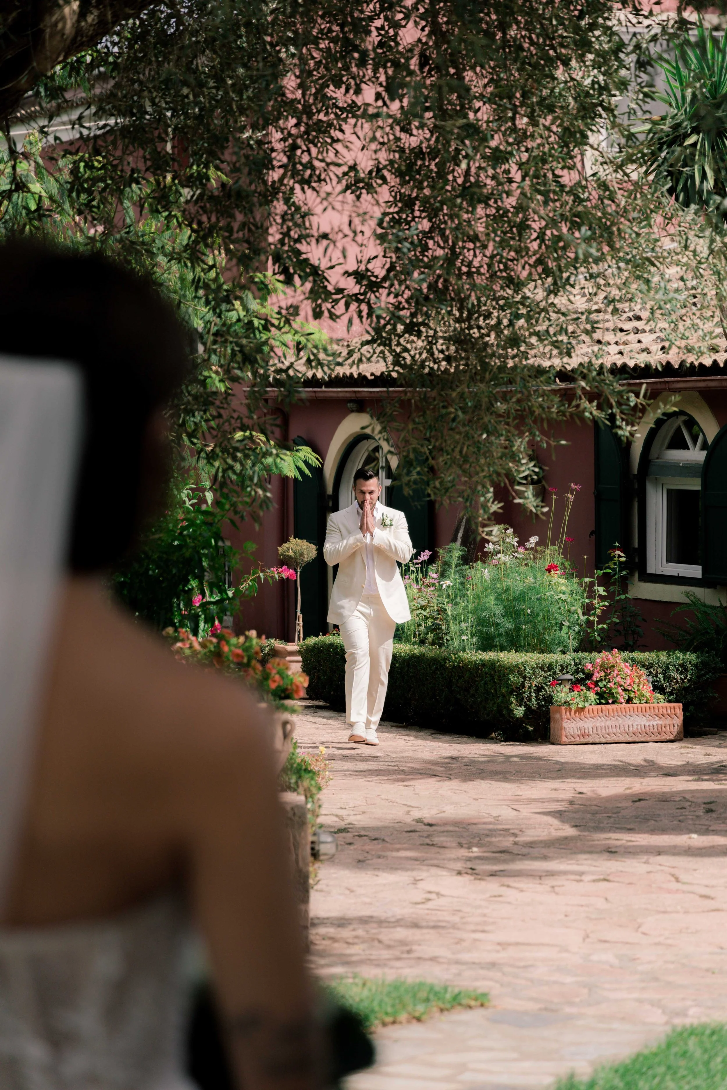 A man dressed in a white suit, walking towards a garden area, with his hands pressed together in a prayer or greeting gesture, seen through blurred figures in foreground, with a pink building, plants, and flowers in the background.