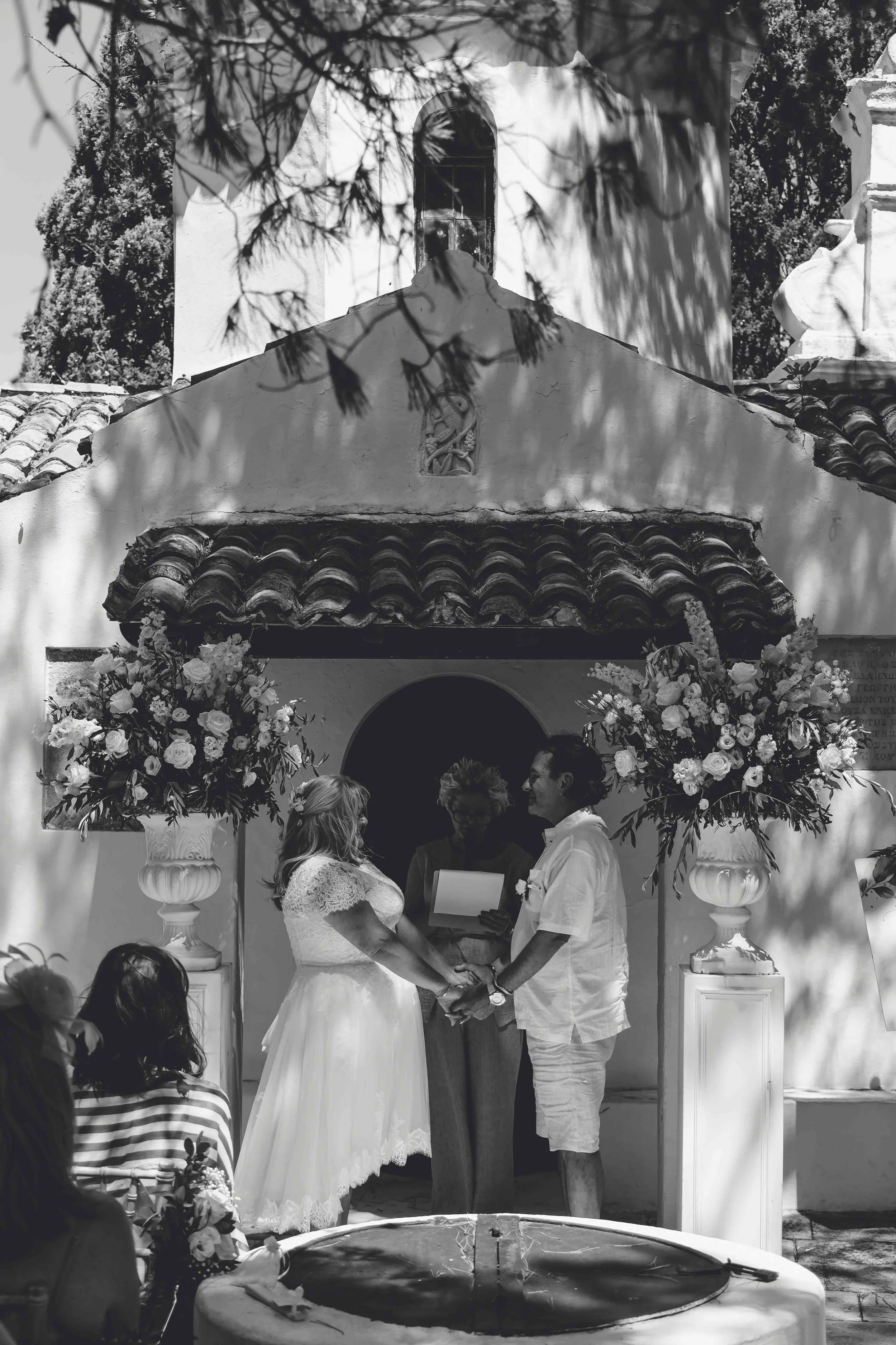 A couple getting married outdoors, holding hands and facing each other, with an officiant standing behind them, during a wedding ceremony. The setting includes large floral arrangements and is under a decorative arch, with a building and trees in the