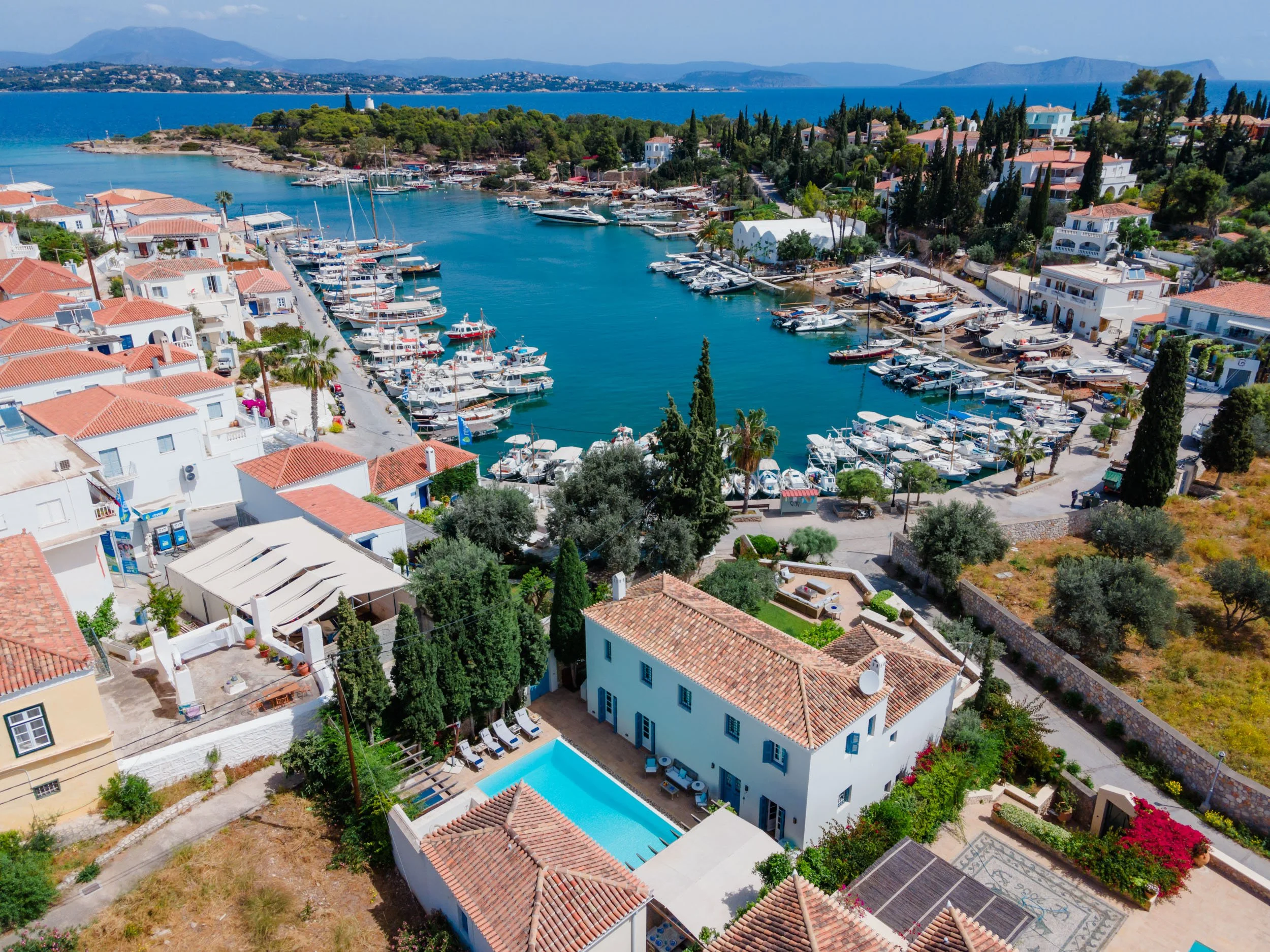 Aerial view of a Mediterranean harbor with boats, surrounded by white buildings with red-tiled roofs, lush greenery, and a swimming pool in the foreground, with mountains in the background.