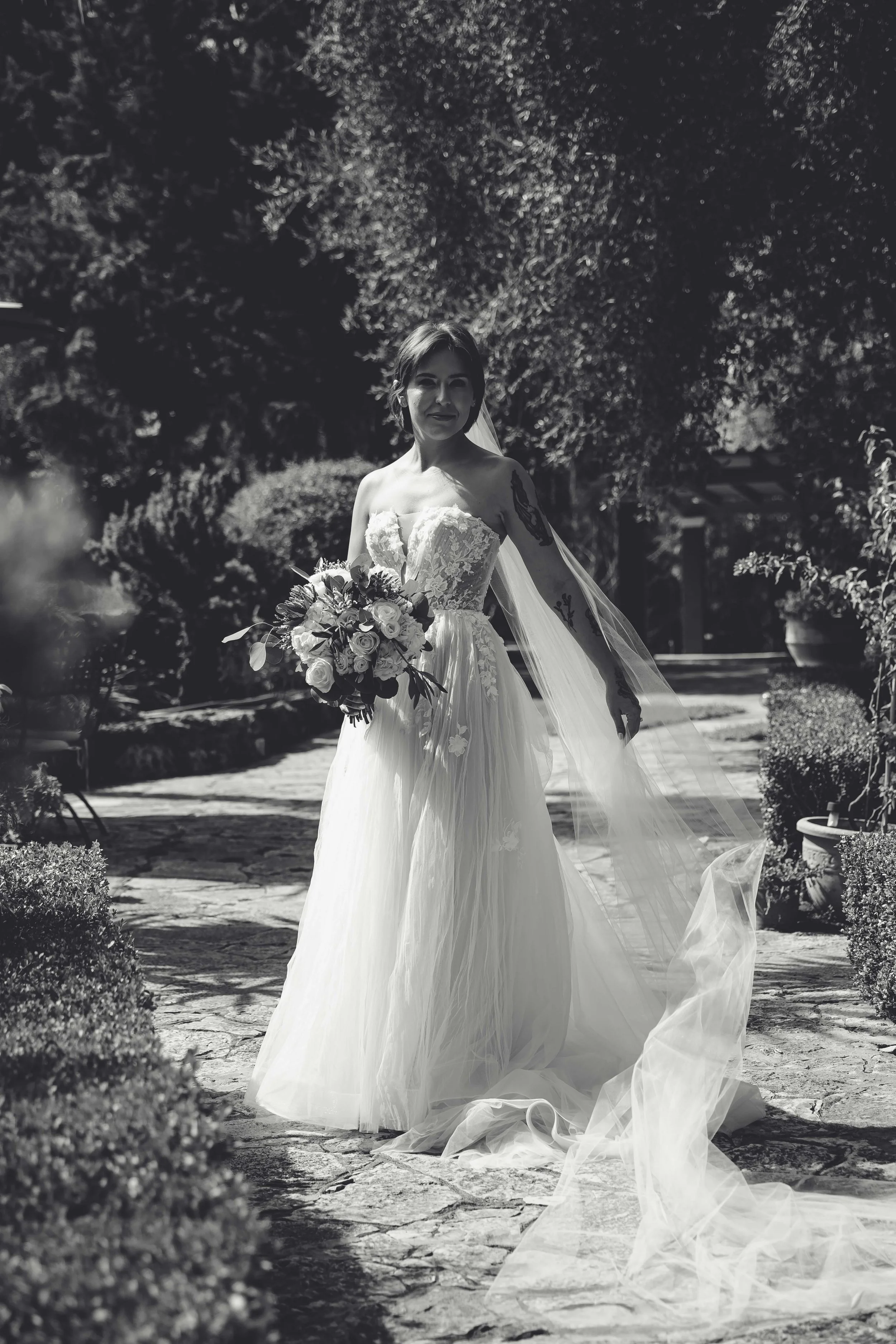 A bride in a wedding dress holding a bouquet, standing outdoors on a stone pathway surrounded by bushes and trees.