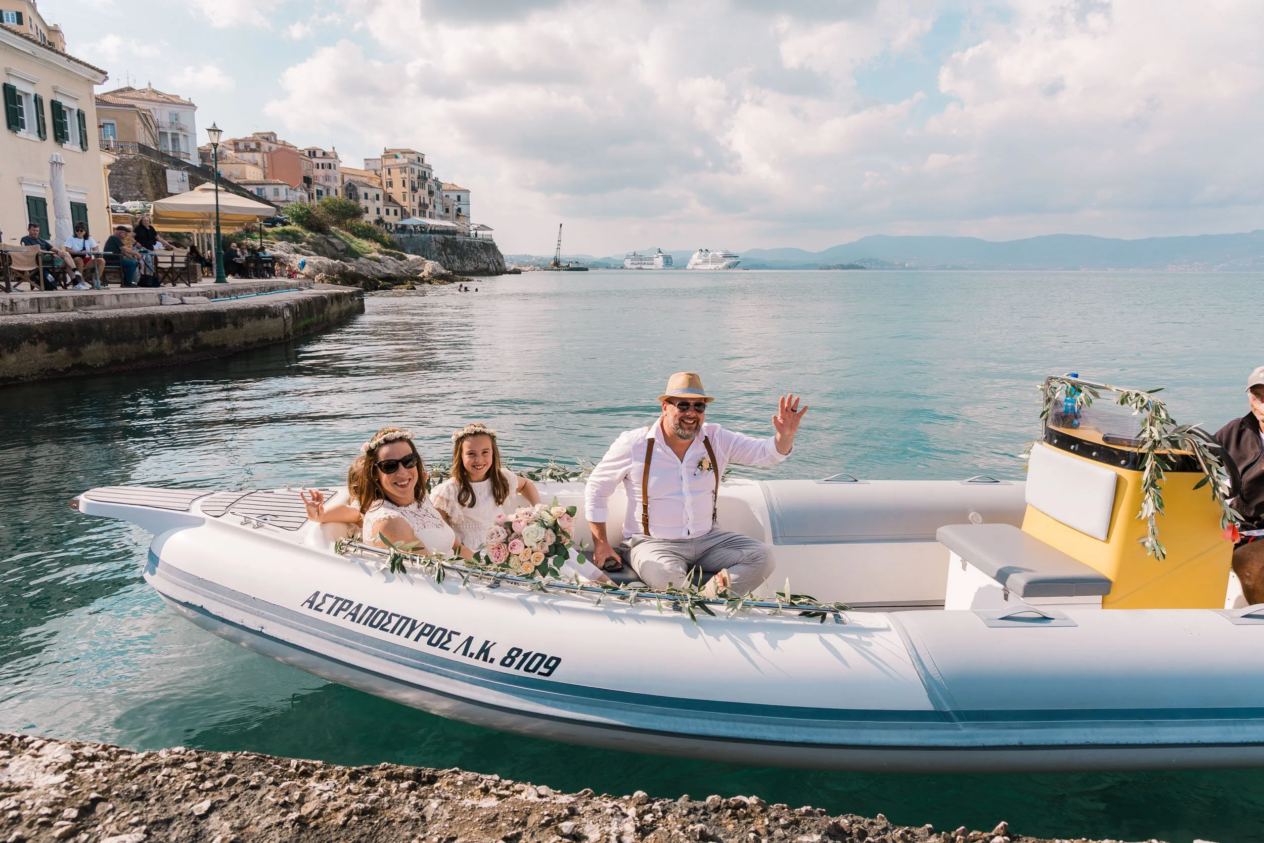 A boat decorated with flowers carrying a bride, a groom, and a man, on a harbor with a seaside town and cruise ships in the background.