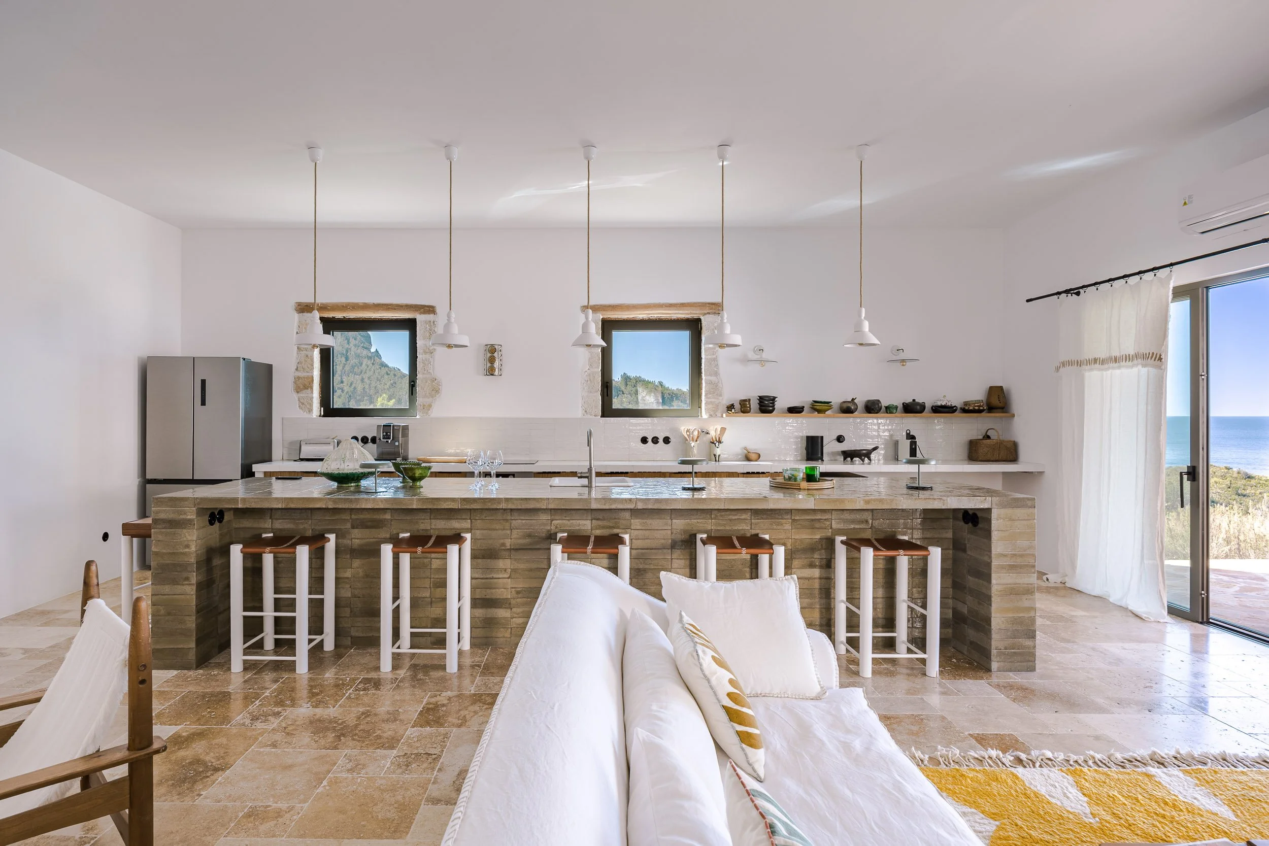 Open-concept kitchen and living room with large island, white walls, beige tiled floor, and sliding glass door leading to outdoor view of ocean and landscape.
