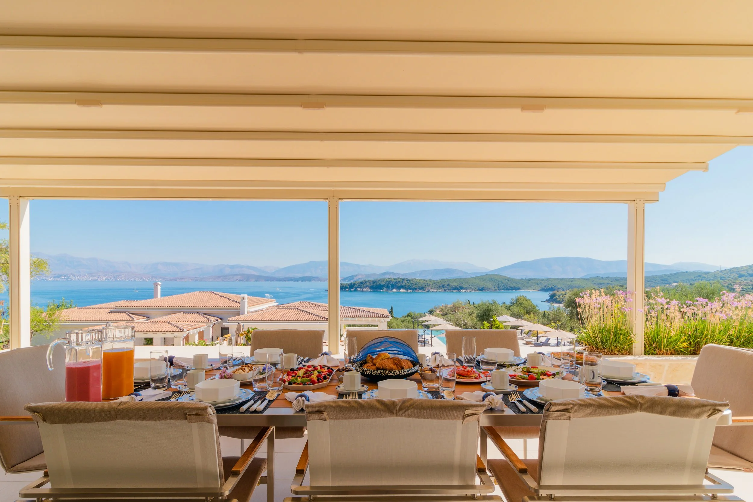A luxurious outdoor dining table set for breakfast with a scenic view of a lake, mountains, and clear blue sky, featuring breakfast foods, drinks, and flowers.