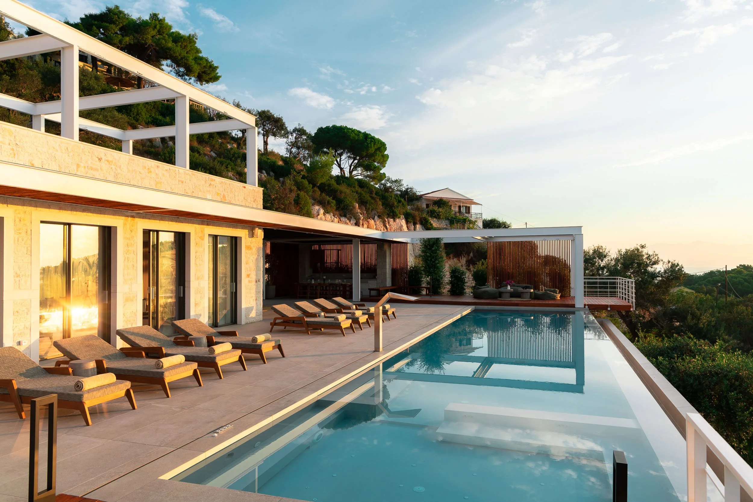 Luxury modern house with an outdoor pool, wooden lounge chairs, and scenic hillside views during sunset.