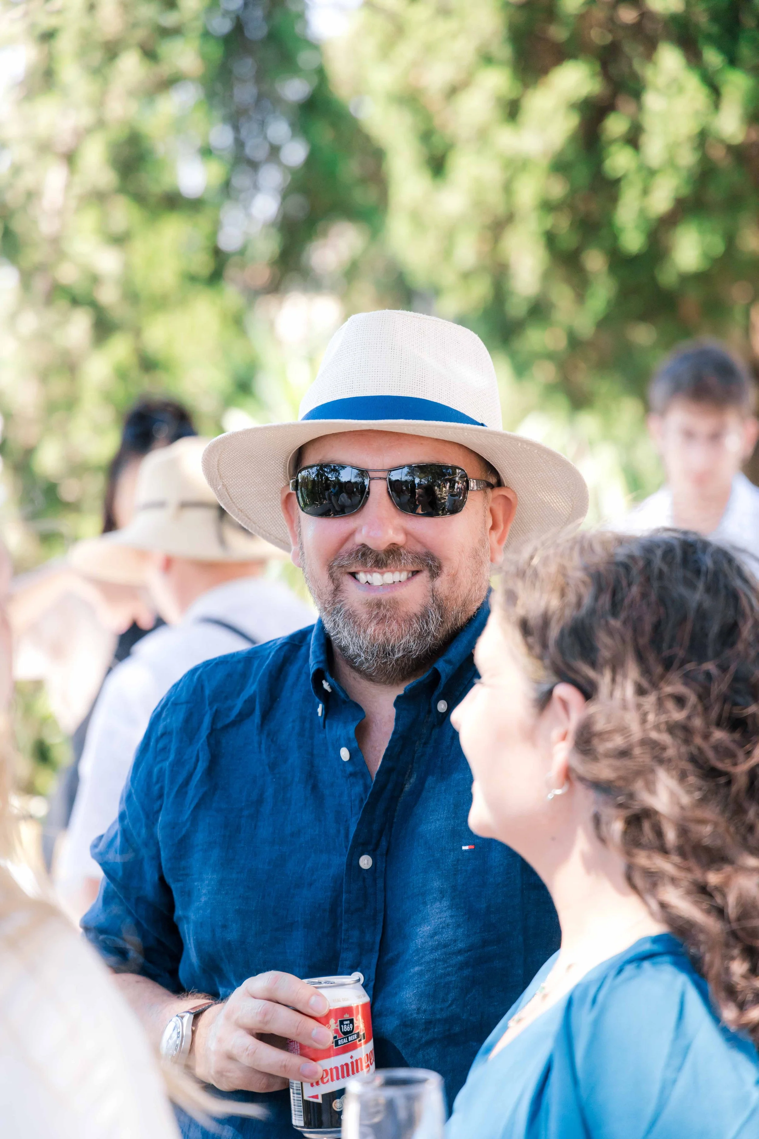 A man with a beard wearing sunglasses, a white fedora, and a blue shirt, smiling and holding a can of beer at an outdoor gathering.