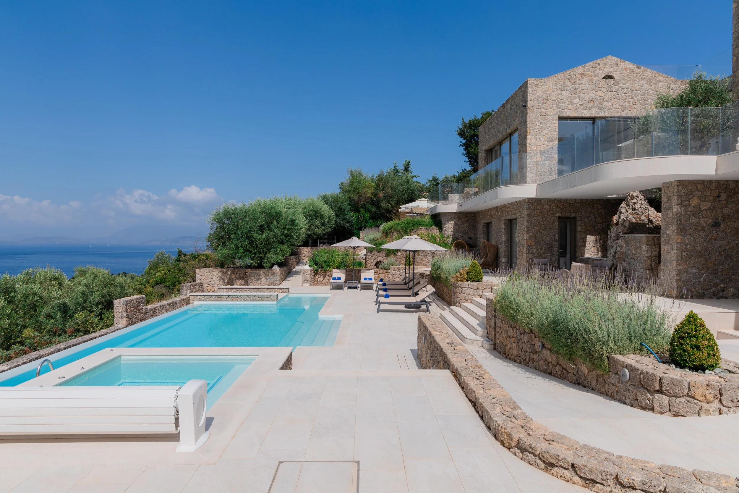 Luxury modern house with stone exterior and glass balcony, outdoor pool with surrounding lounge chairs and umbrellas, landscaped garden with bushes and trees, overlooking a scenic ocean view under clear blue sky.