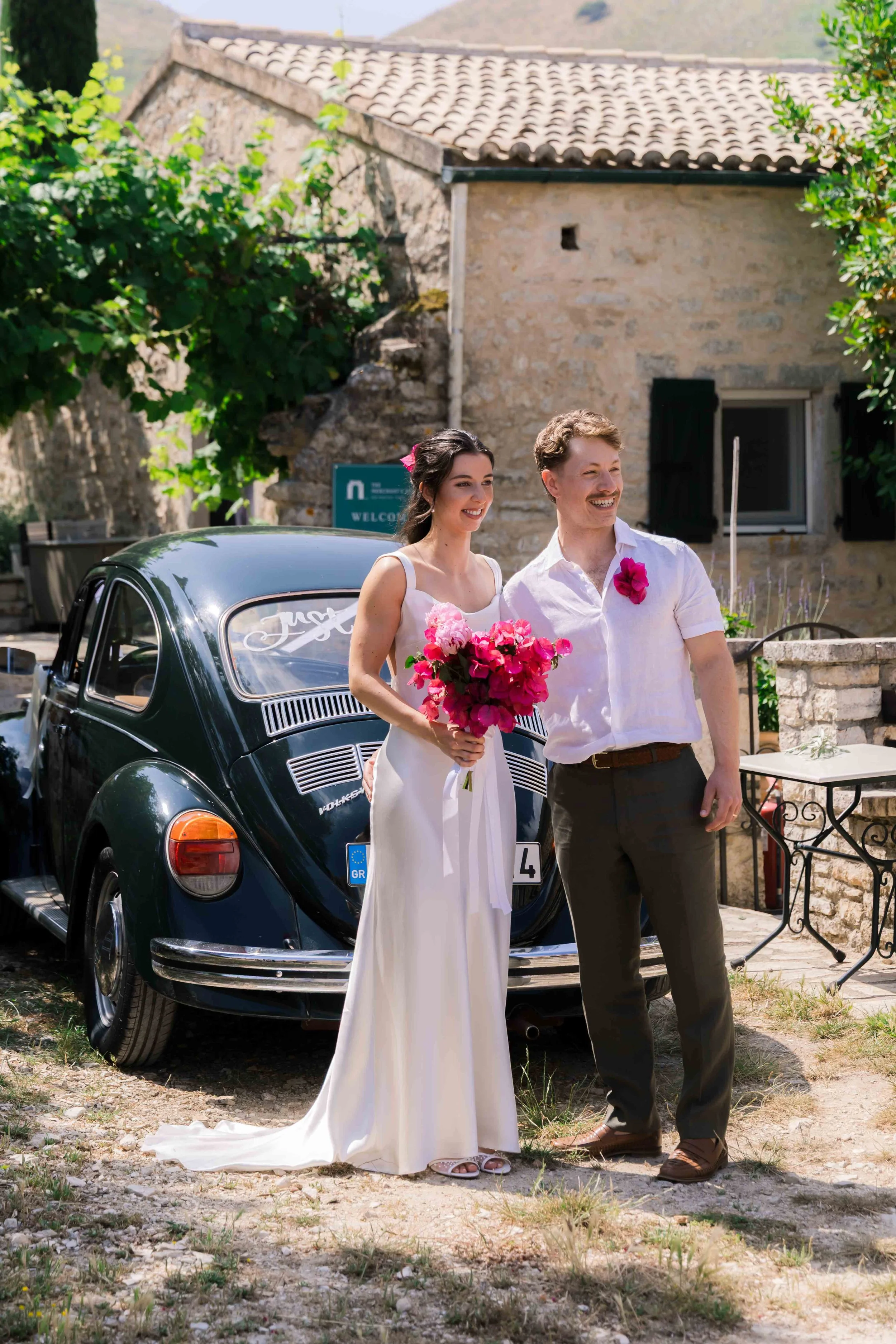 A bride in a white wedding dress holding a bouquet of pink flowers standing next to a groom in a white shirt with a pink flower pinned to his shirt, both smiling outdoors near a vintage black car with a European license plate, in front of a rustic st