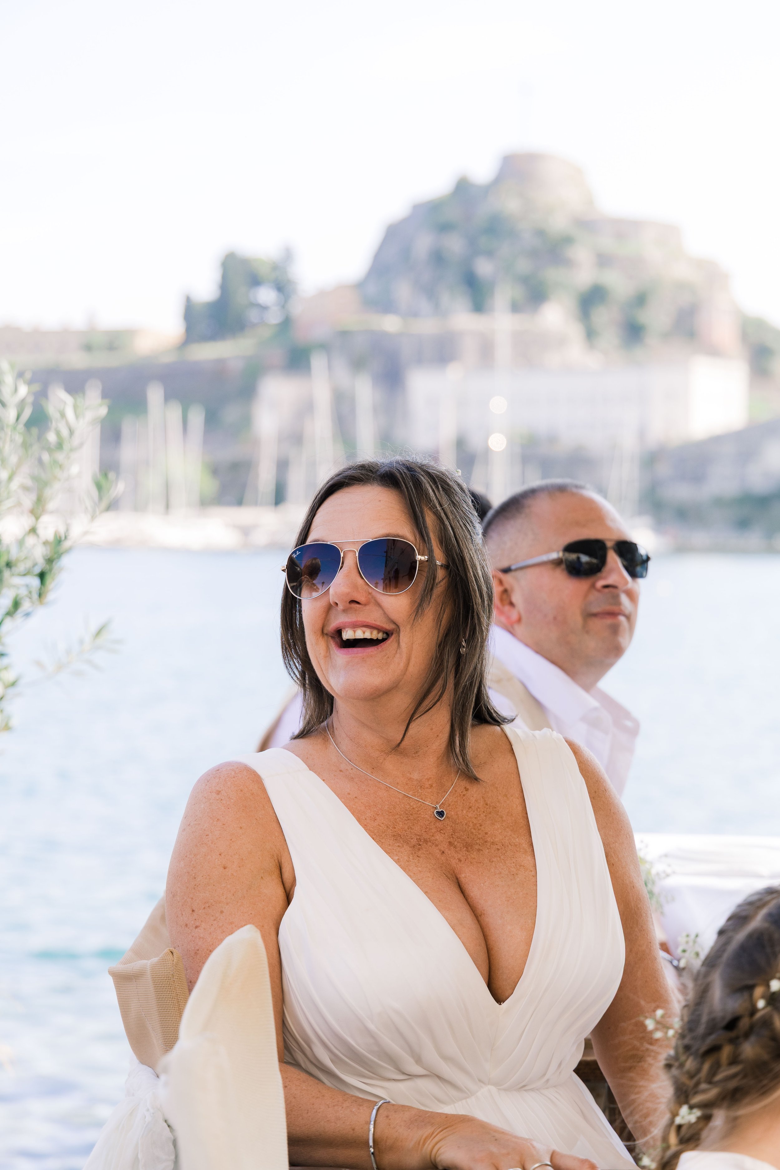 Woman with dark hair and sunglasses smiling at an outdoor event near water, with a man in dark sunglasses behind her, and boats and a hillside in the background.